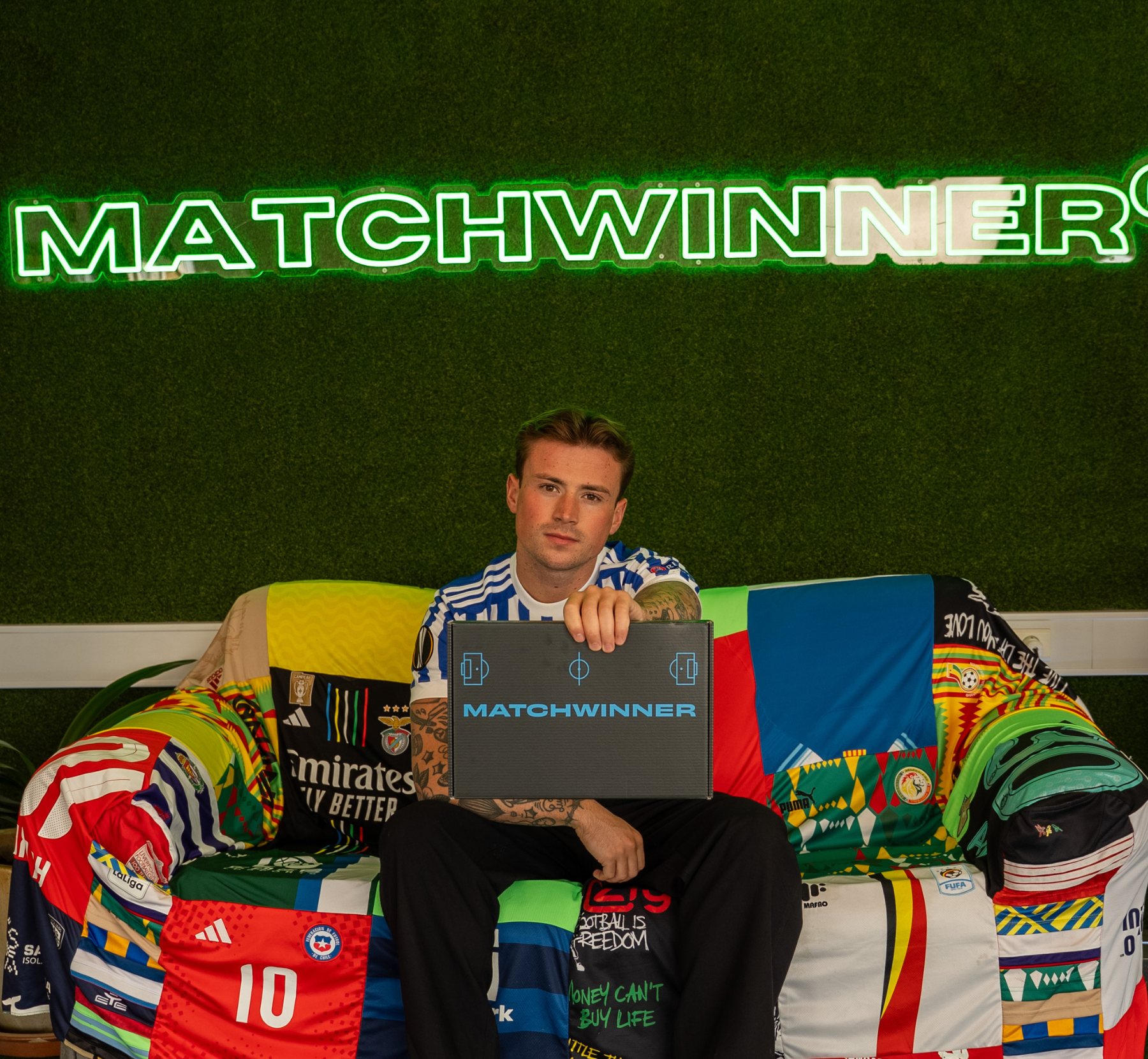 A man sits on a couch made of football jerseys, holding a black box under a neon sign.