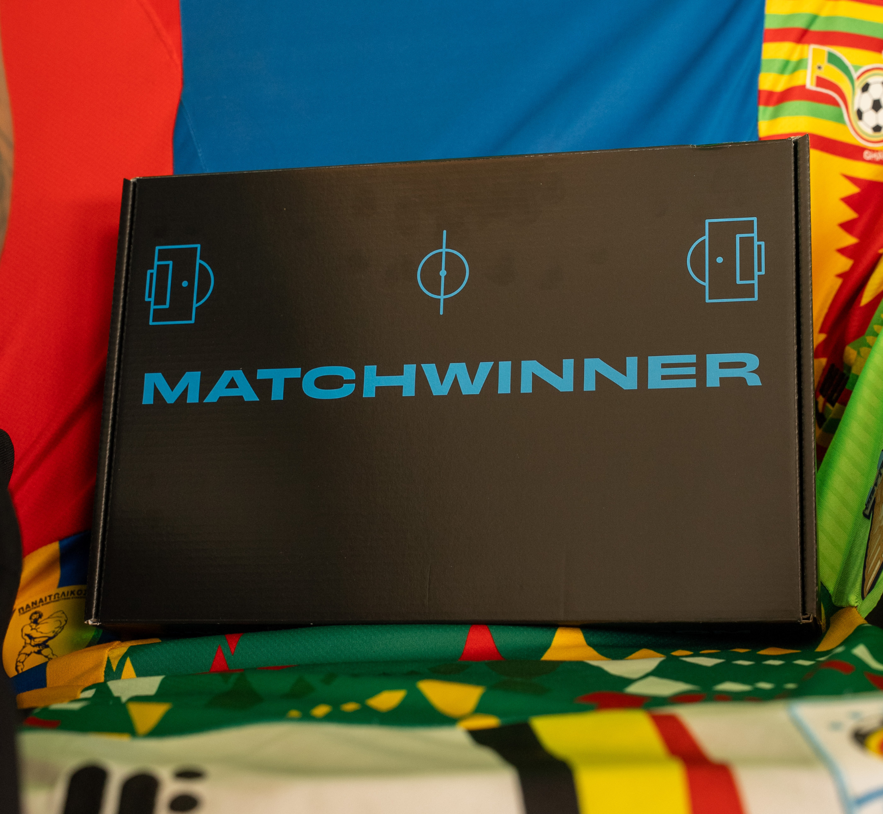 A black box with the word MATCHWINNER in blue text, resting on a pile of colorful jerseys.