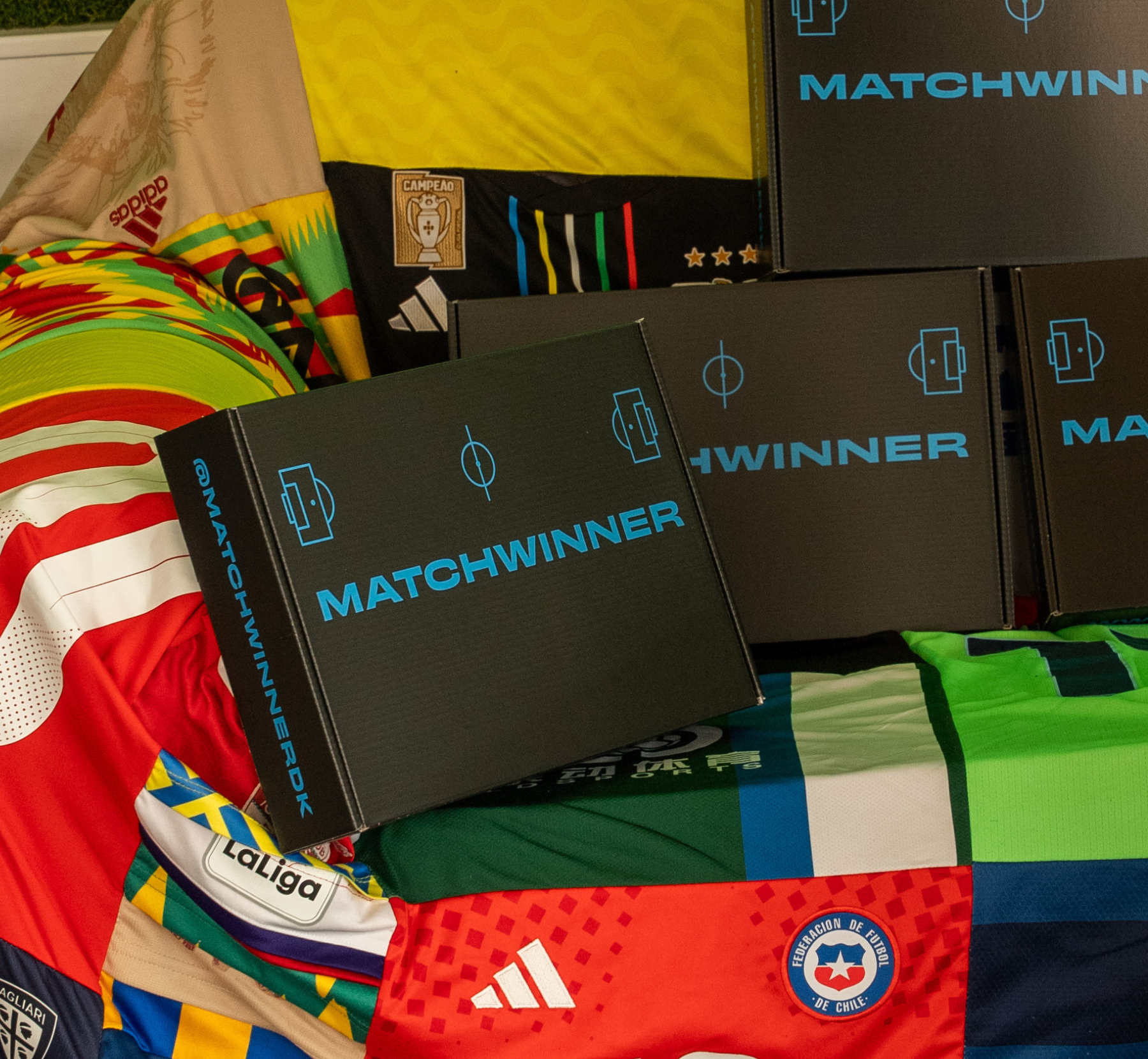 Several black Matchwinner boxes sitting on top of a colorful background made of various soccer jerseys.