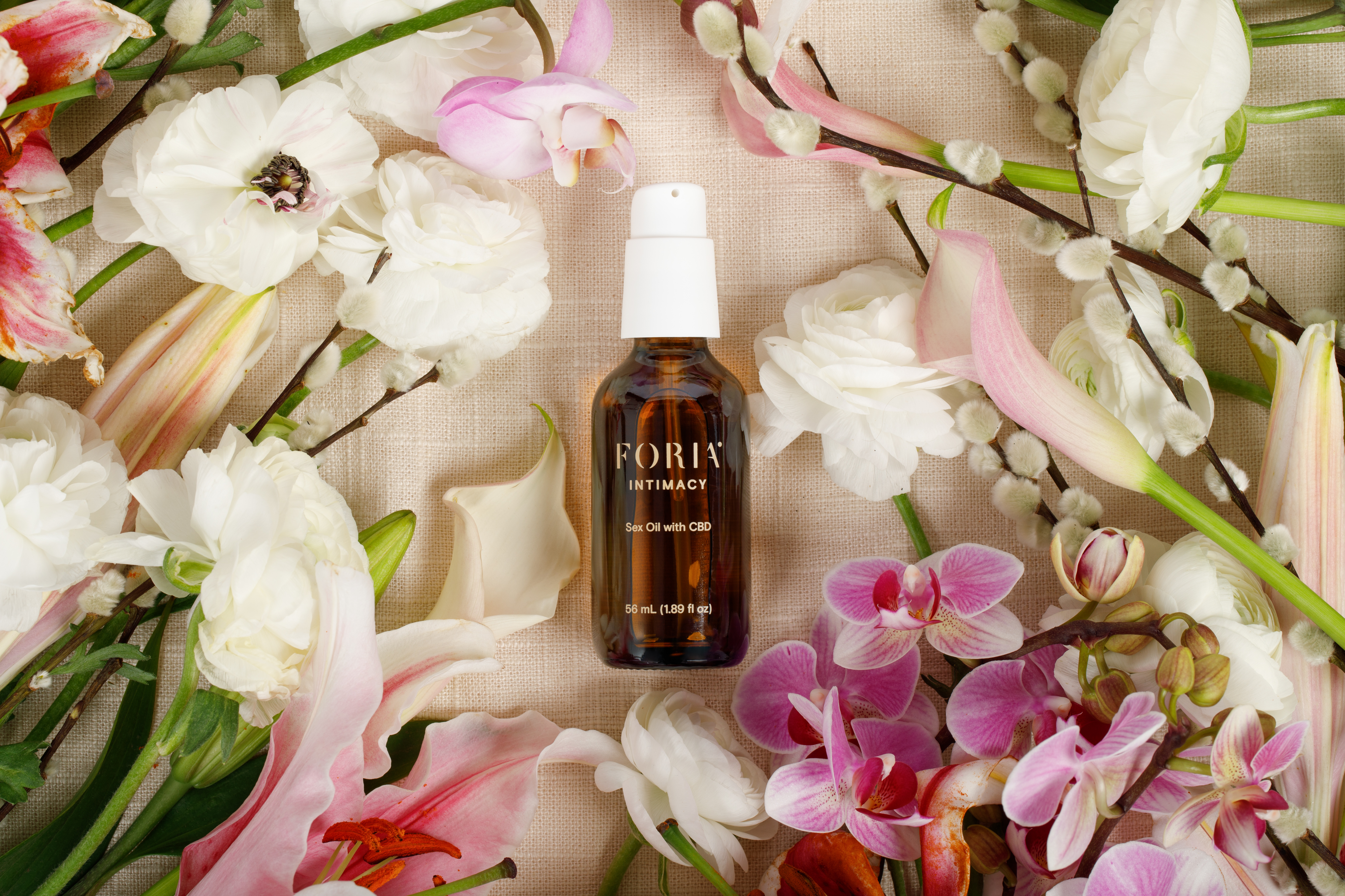 A brown bottle labeled 'Foria Intimacy' surrounded by various flowers on a fabric surface.