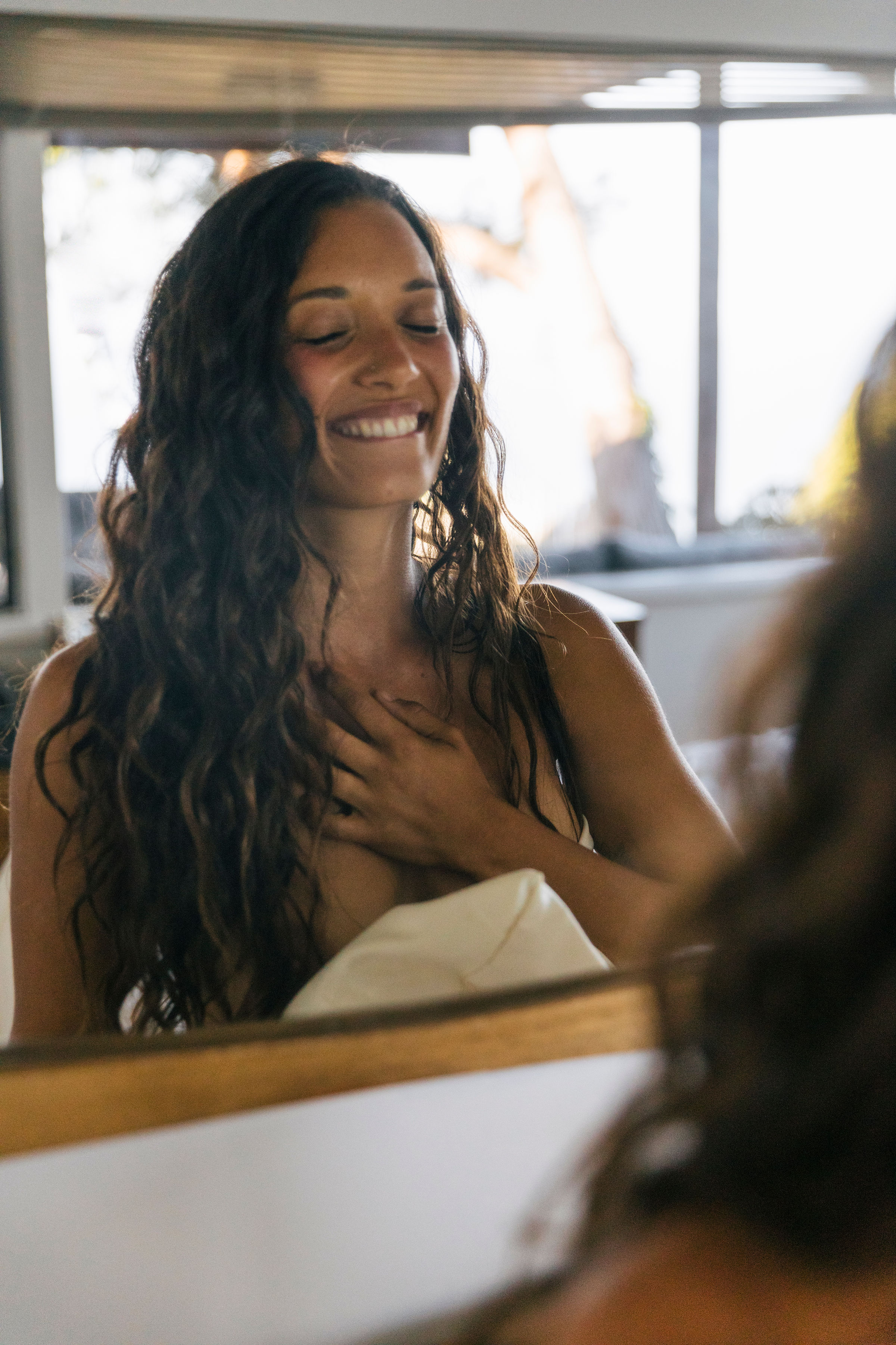 Smiling woman with long curly hair, hand on chest, indoors.