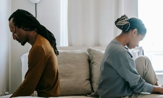 Two people sitting back-to-back on a bed, looking down.
