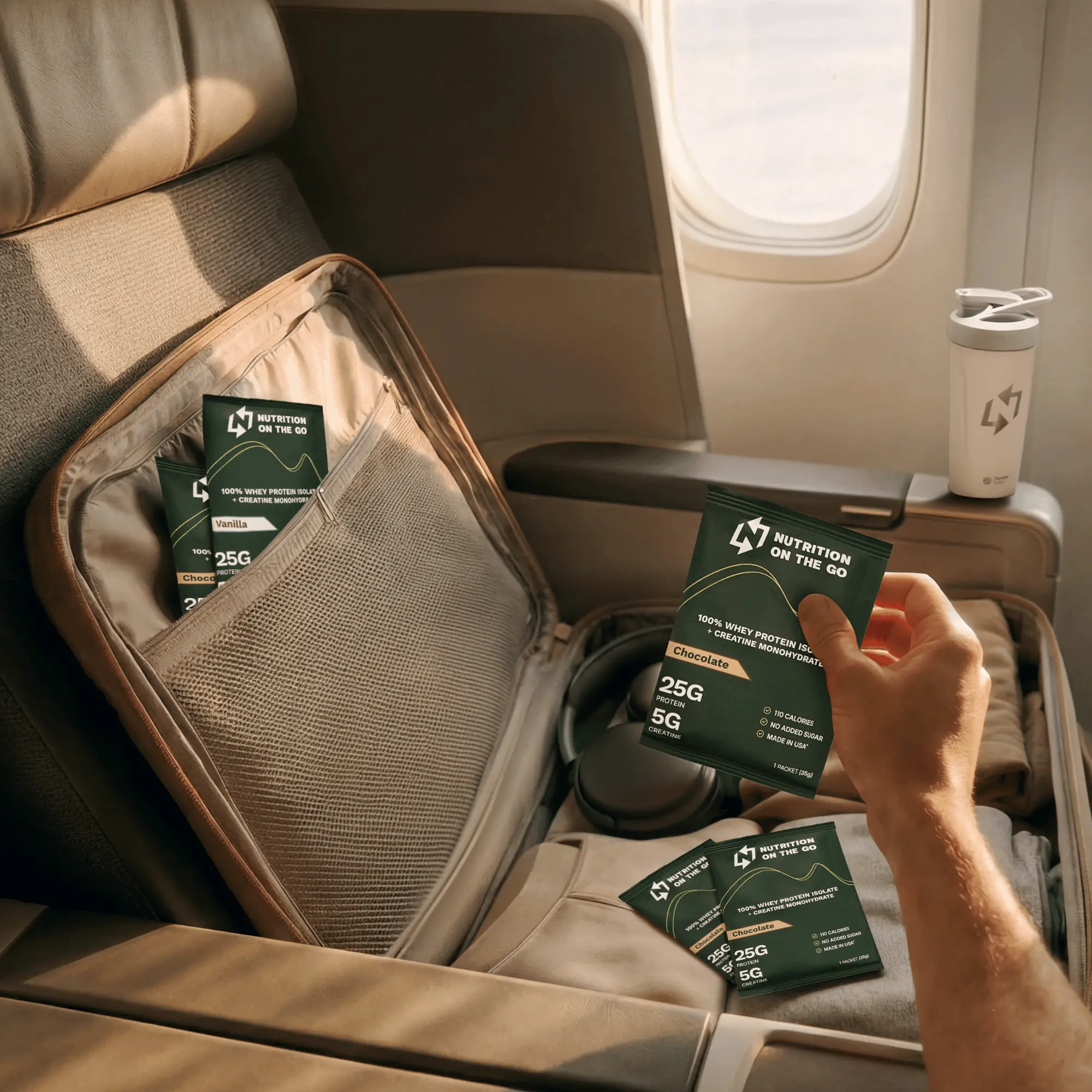 A hand holding a single-serve packet of protein powder on a plane. They are traveling and there is a suitcase with clothes, and other packets on a seat in first class.