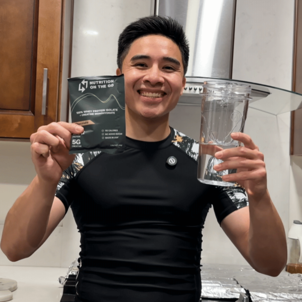 A smiling man in a kitchen holds a protein powder packet and a shaker bottle.