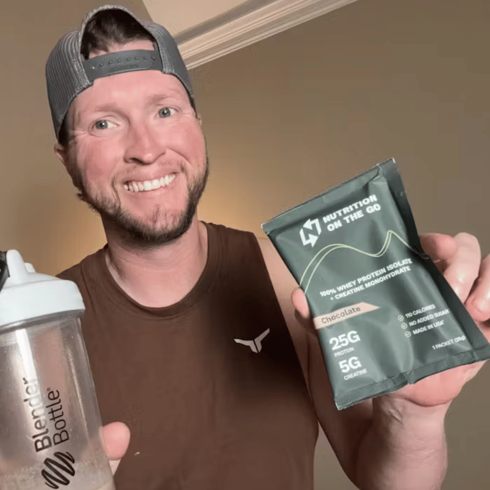 A man smiling and holding a single serve packet of whey protein + creatine and a blender bottle.