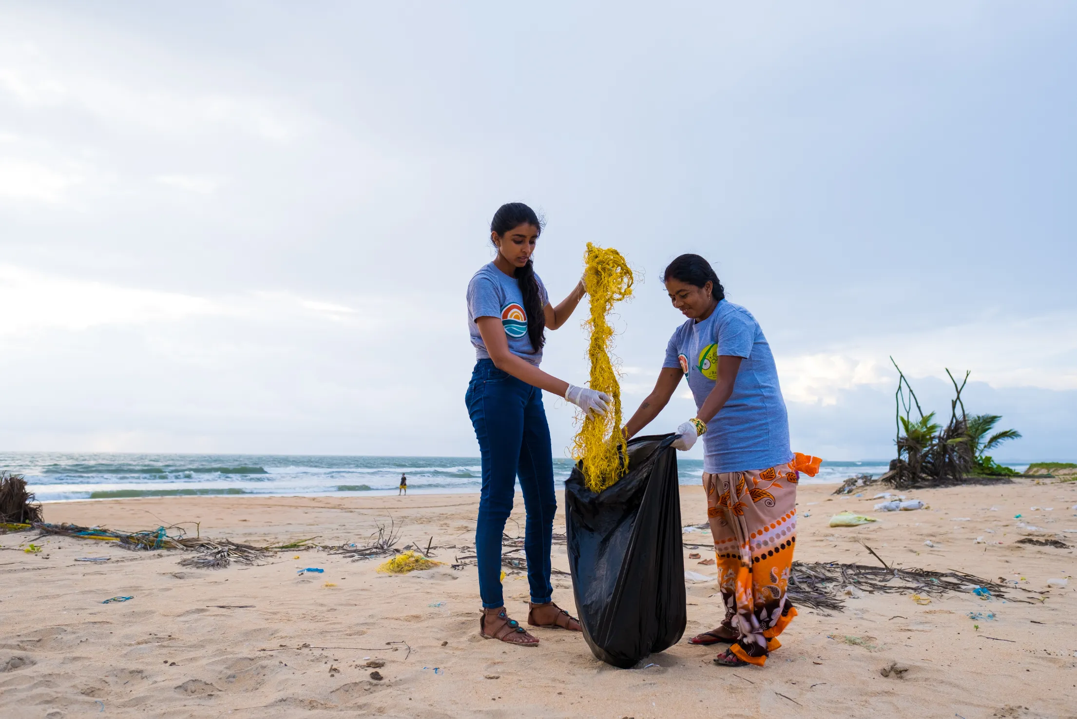 Two people collecting plastic into a bin bag on a litter-strewn beach in Goa, India.