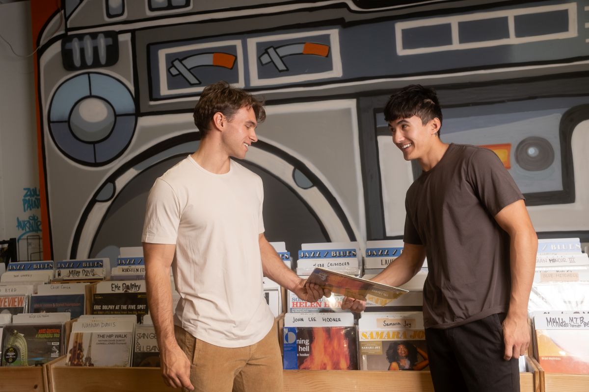 Two men smile at each other while one hands the other a vinyl record in a record store.