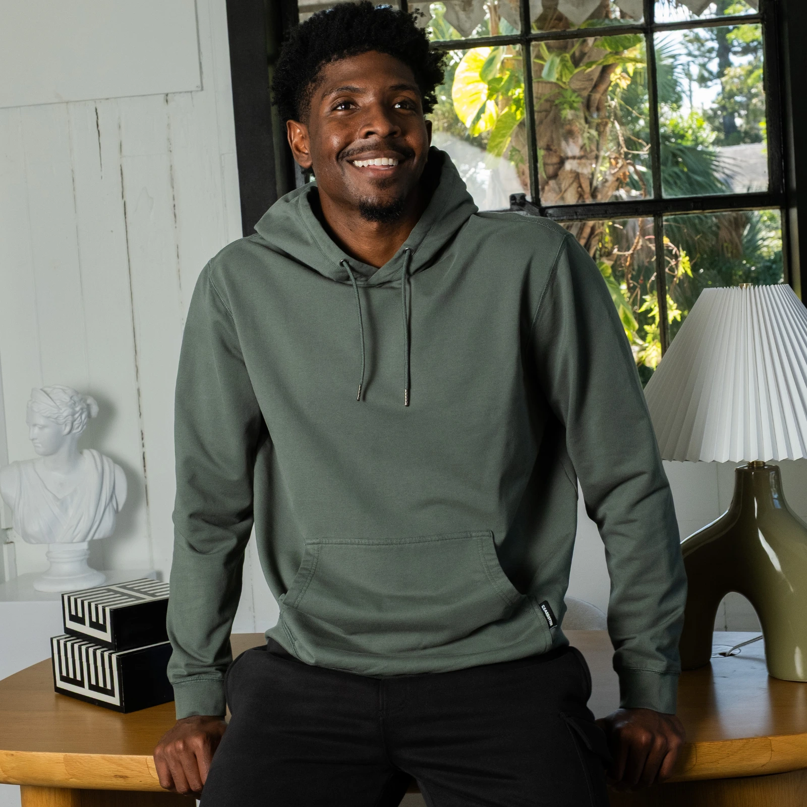 A man in a green hoodie and black pants smiles while sitting on a wooden table indoors.