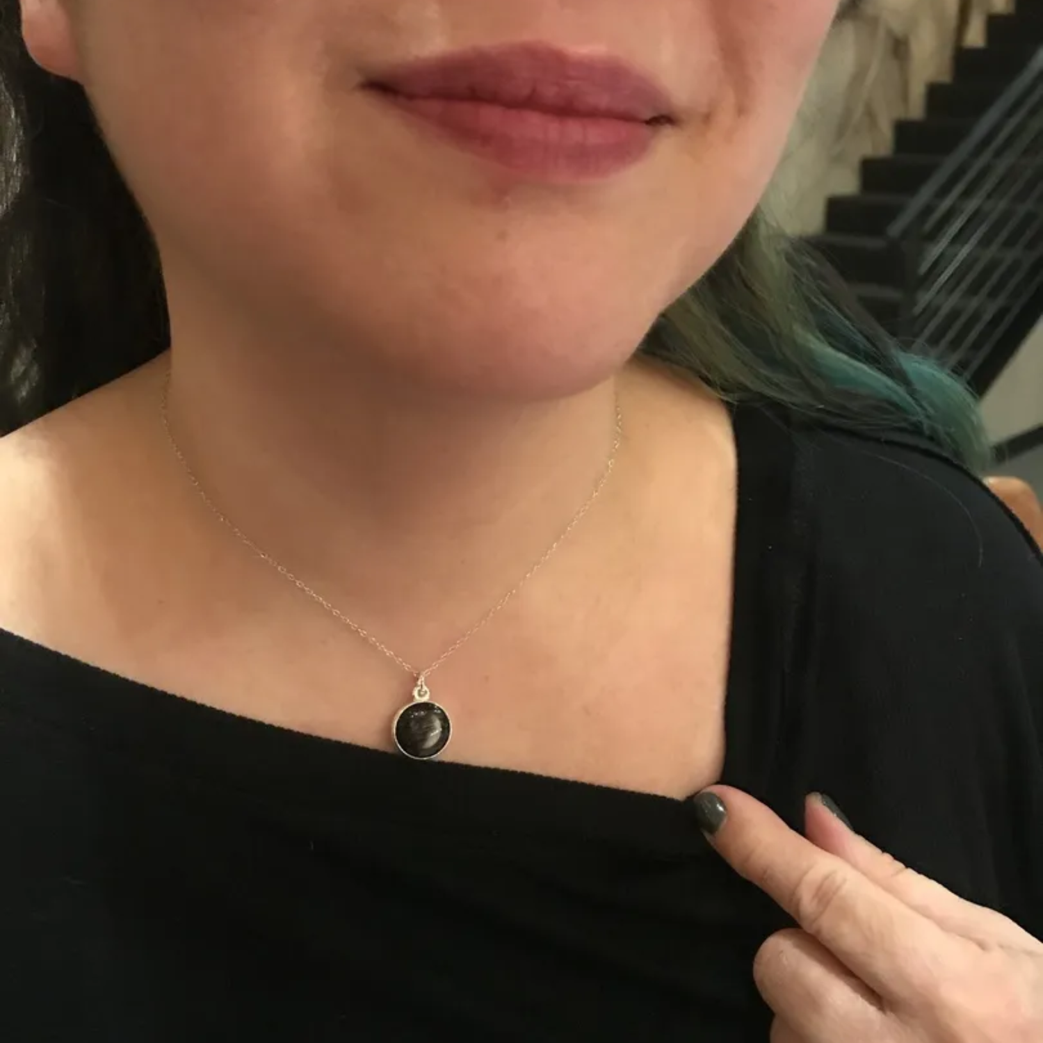 Person wearing a necklace with a round pendant, pointing at it.