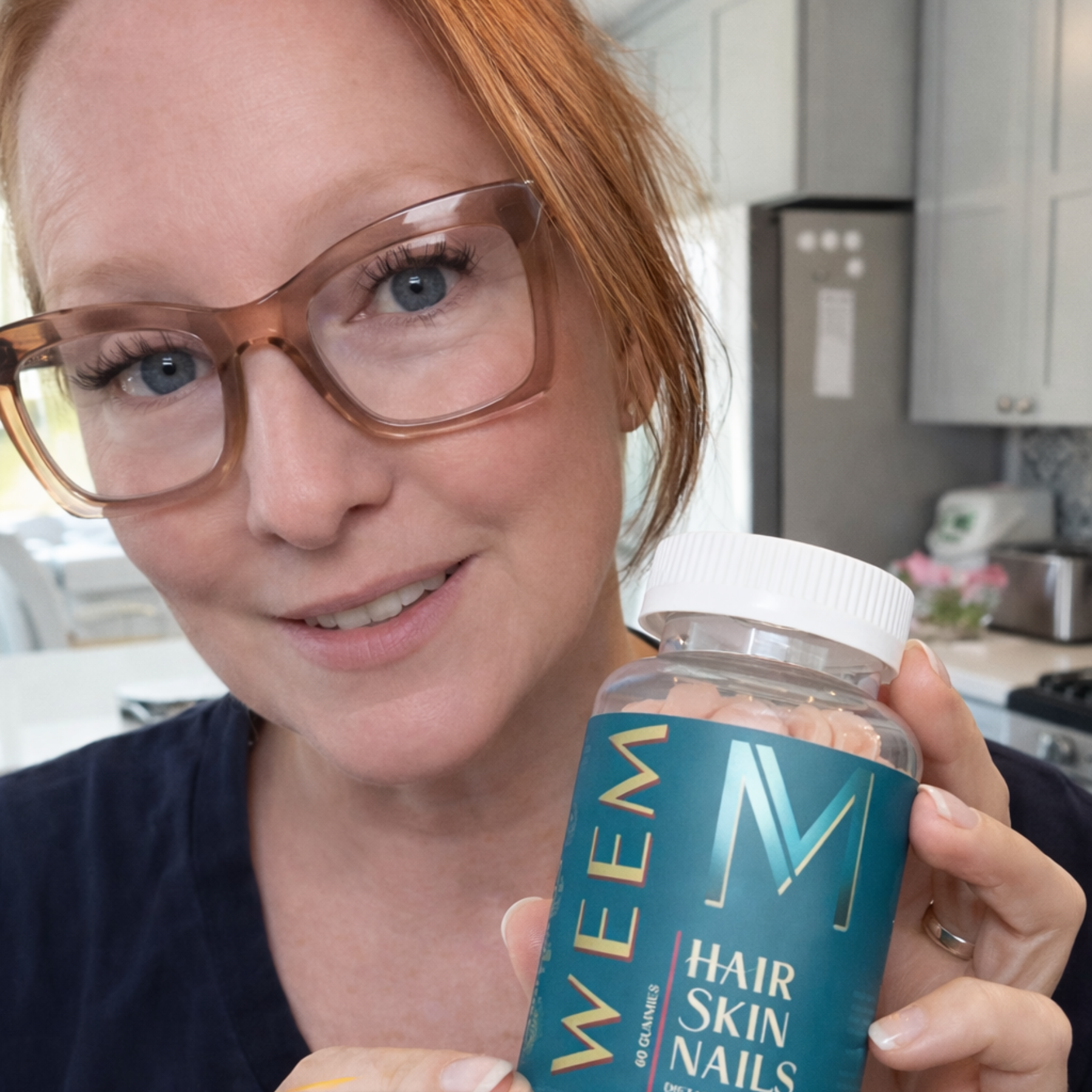 A smiling woman with red hair and glasses holds up a bottle of Weem Hair Skin Nails gummies.