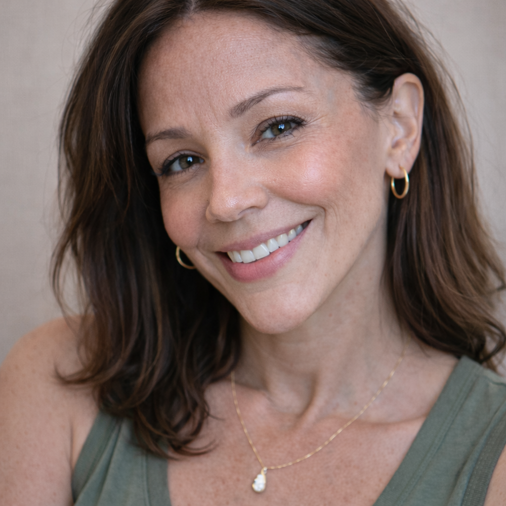 A smiling woman with shoulder-length brown hair, wearing a green tank top and gold jewelry, looks at the camera.