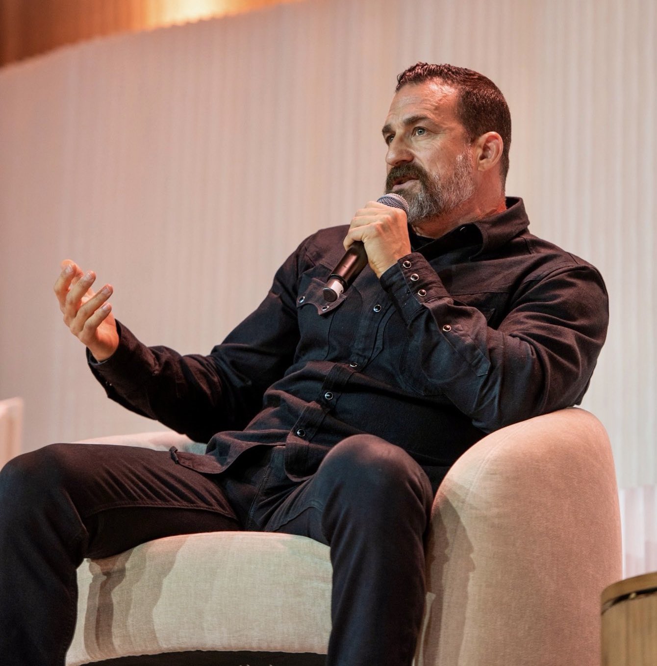 A man with a beard, wearing a black shirt, sits in an armchair speaking into a microphone.