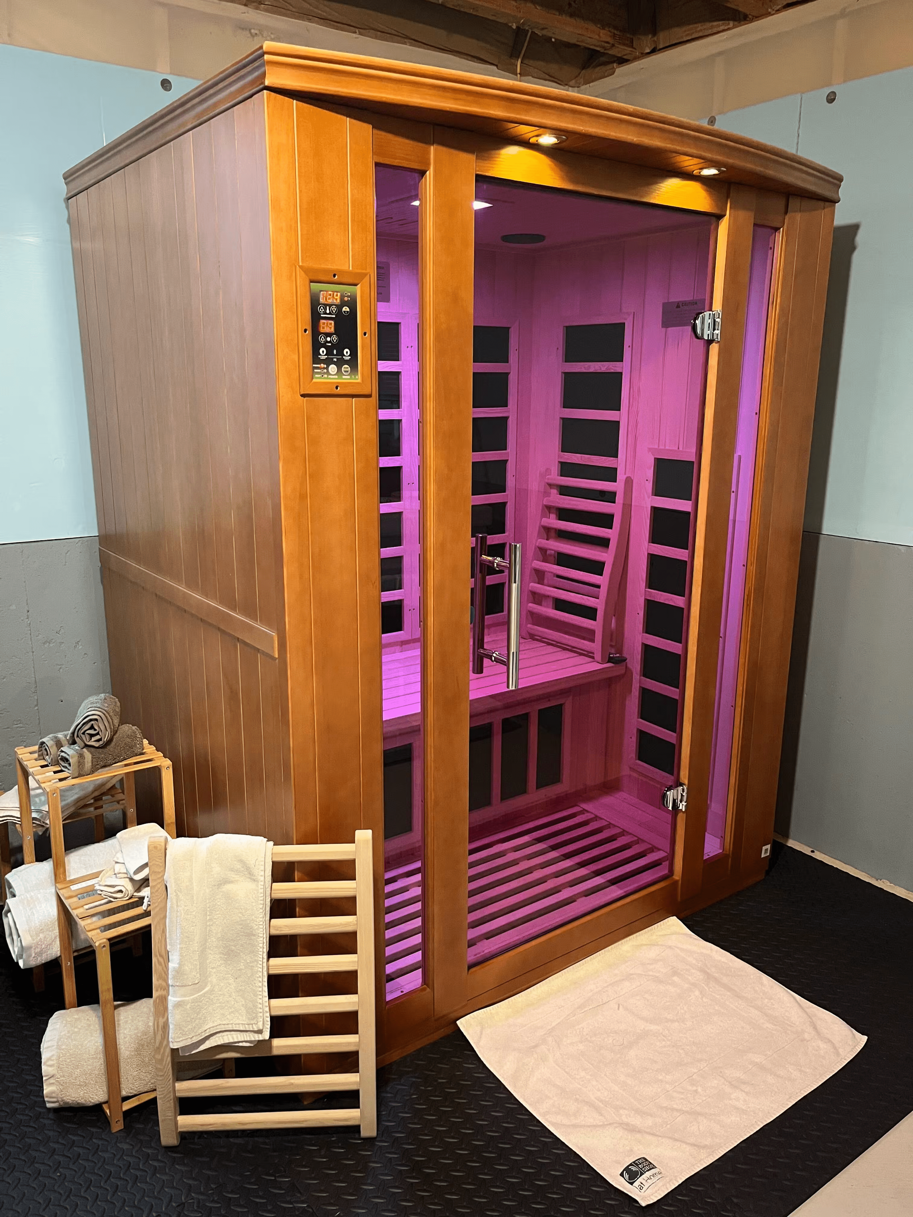 Wooden sauna with glass door, pink interior lighting, and towels on racks nearby.