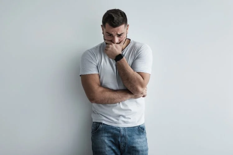 Man in white t-shirt and jeans looking thoughtful against a plain background.