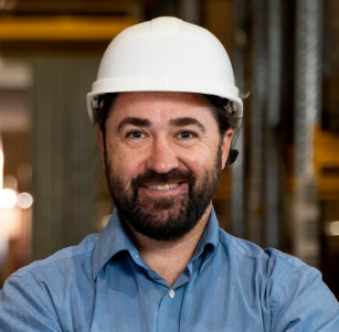 Man in a white hard hat and blue shirt smiling in an industrial setting.
