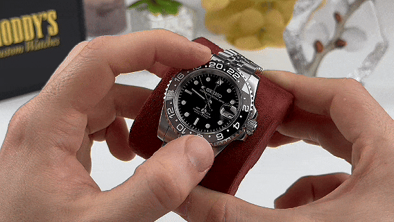 Hands holding a silver watch with a black dial on a cushion.