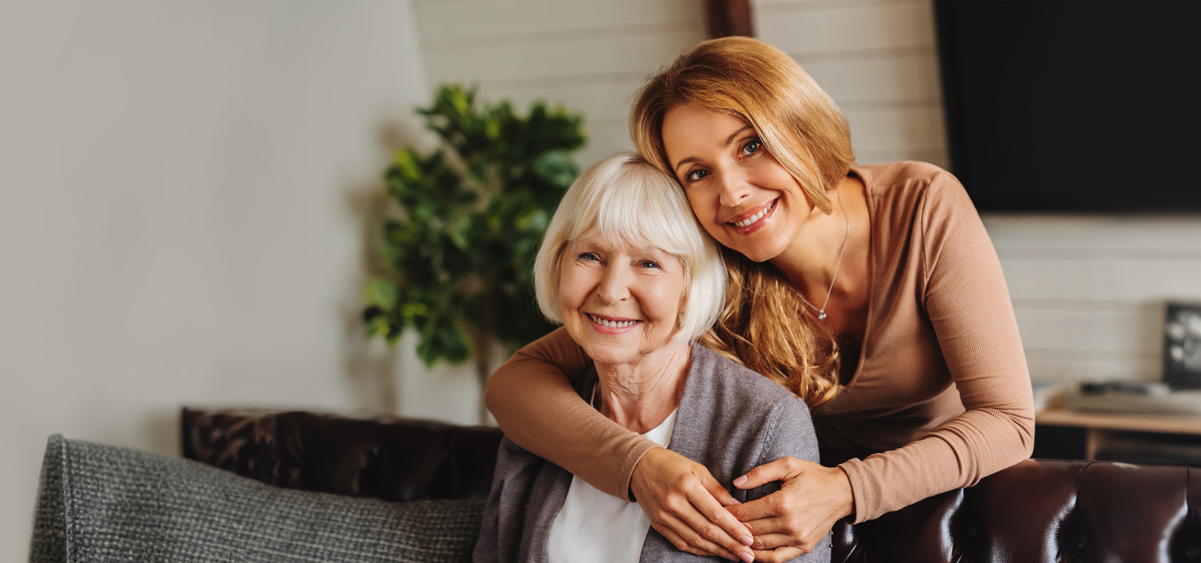 Two women smiling, one embracing the other, indoors with a plant in the background.