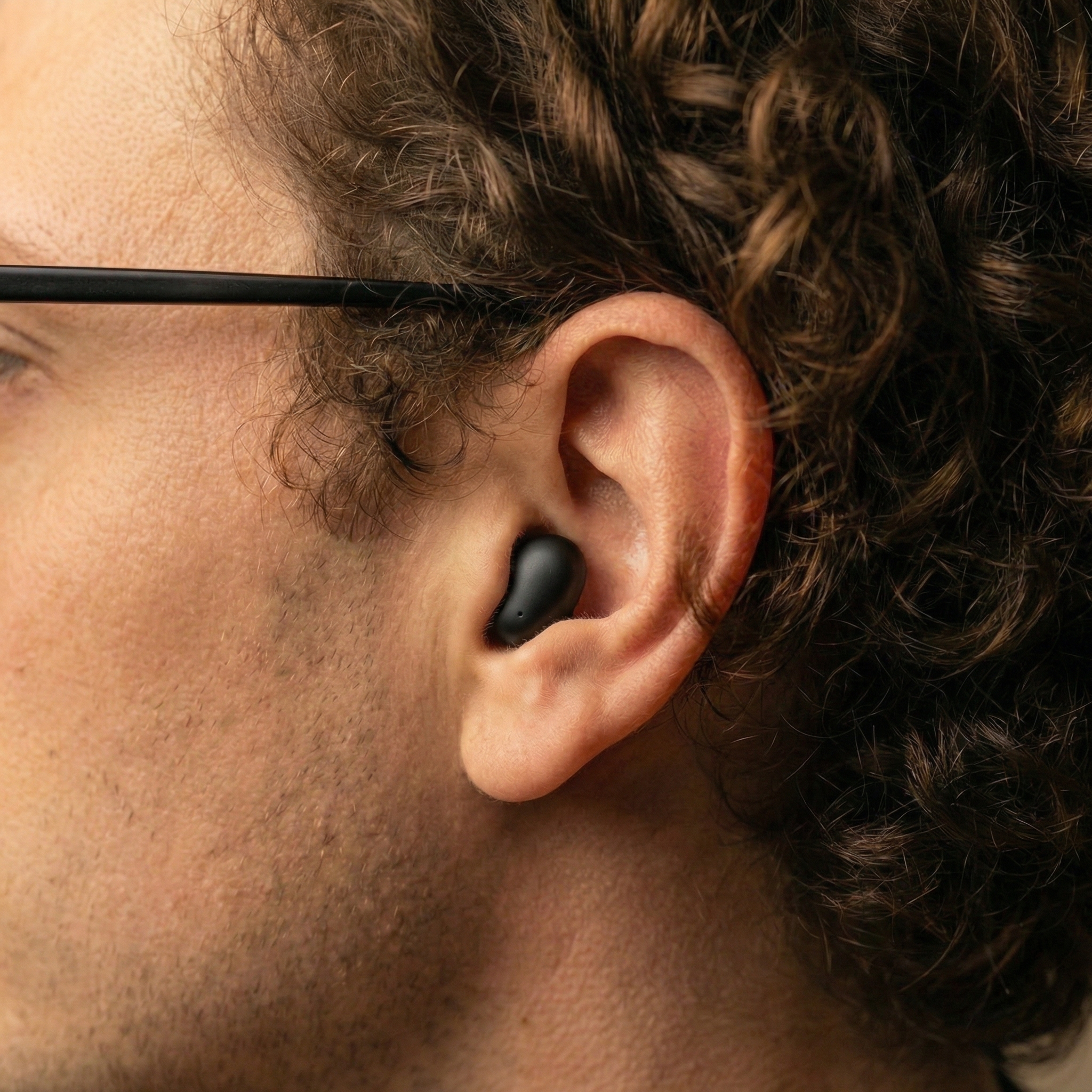 A close-up of a person's ear, showing a black wireless earbud and the arm of their glasses.