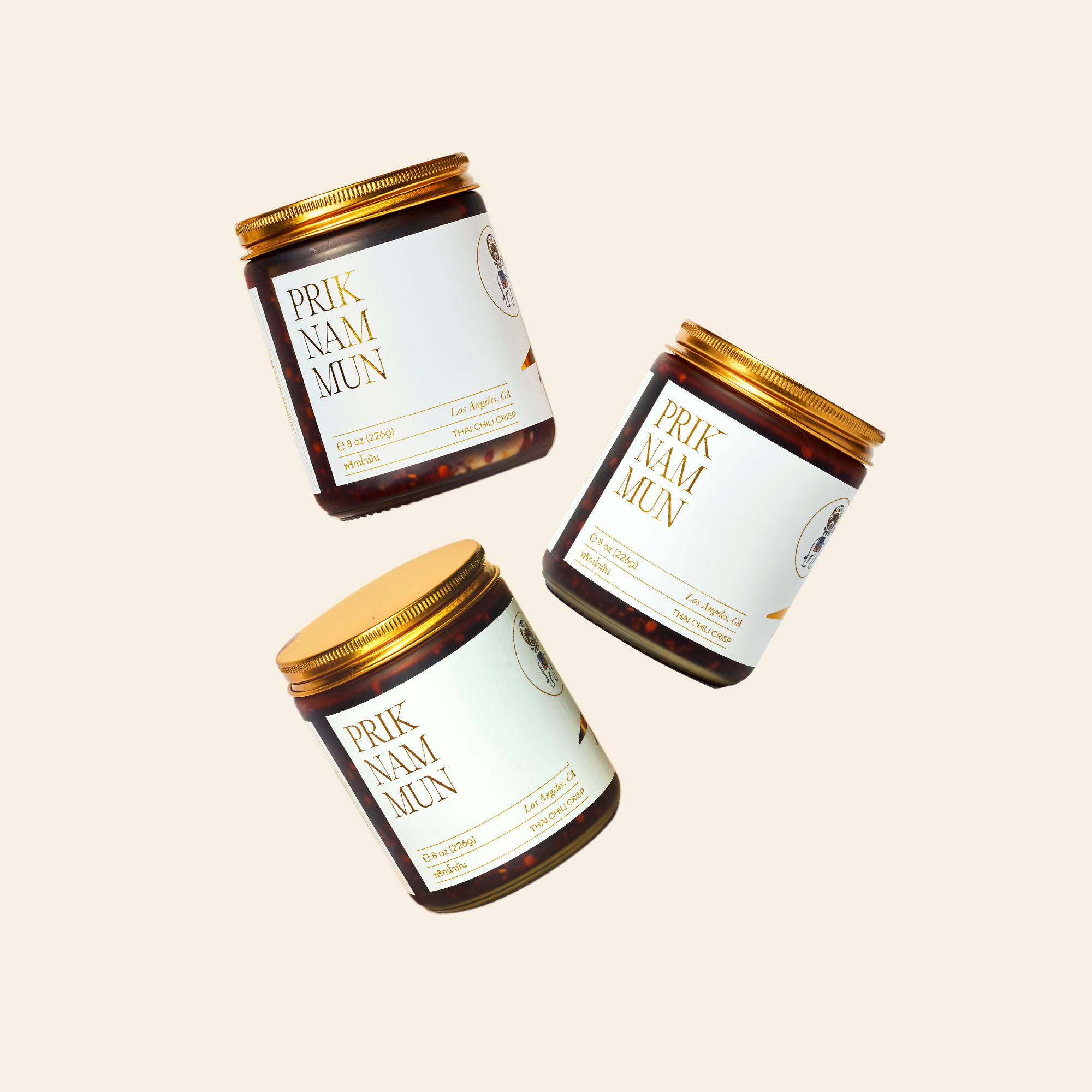 Three jars of Prik Nam Mun Thai Chili Crisp with gold lids floating against a cream background.