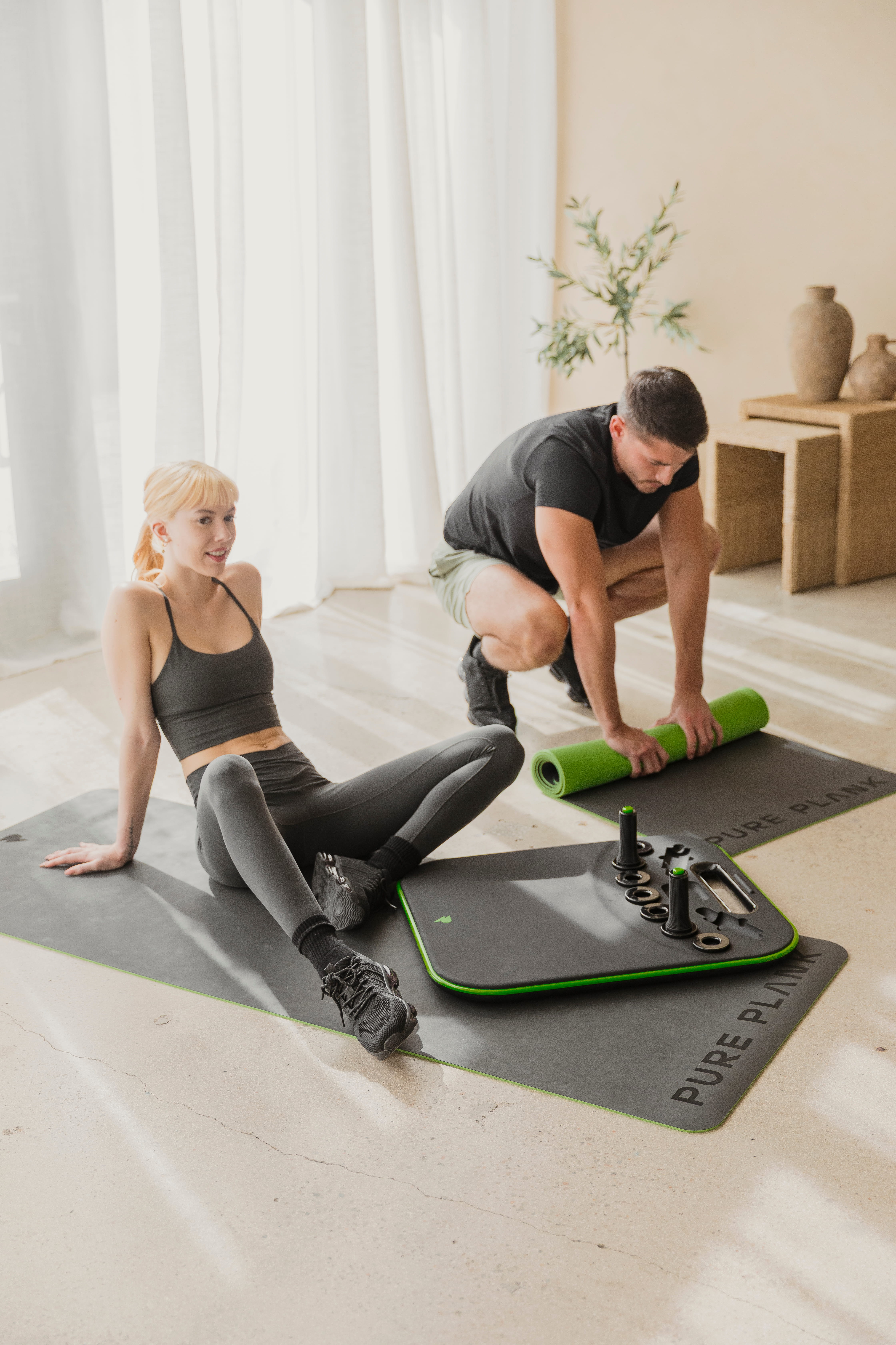 A woman sits on a workout mat while a man rolls up another one next to her.