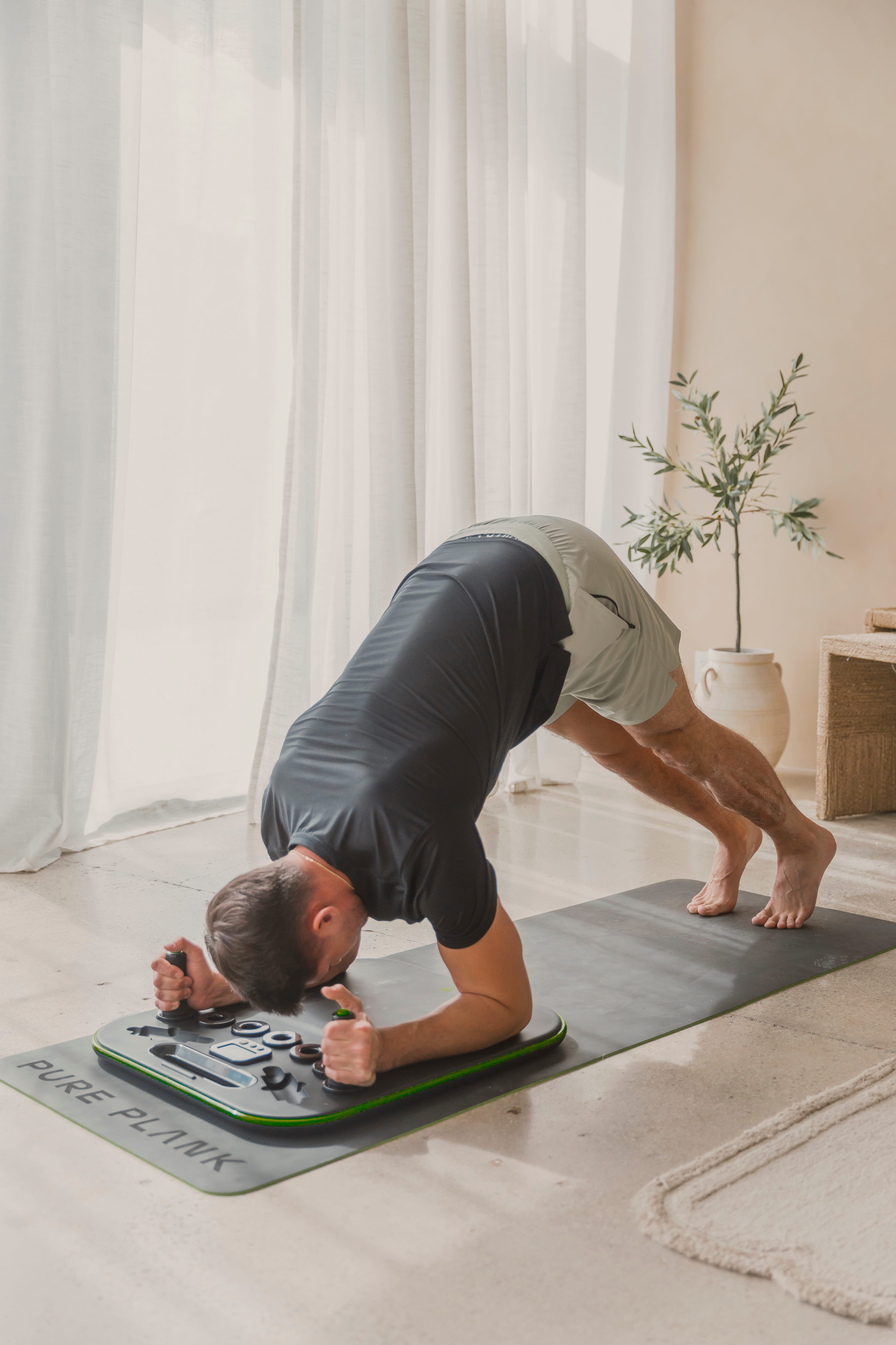 A man in a dolphin pose uses a Pure Plank exercise board on a yoga mat indoors.