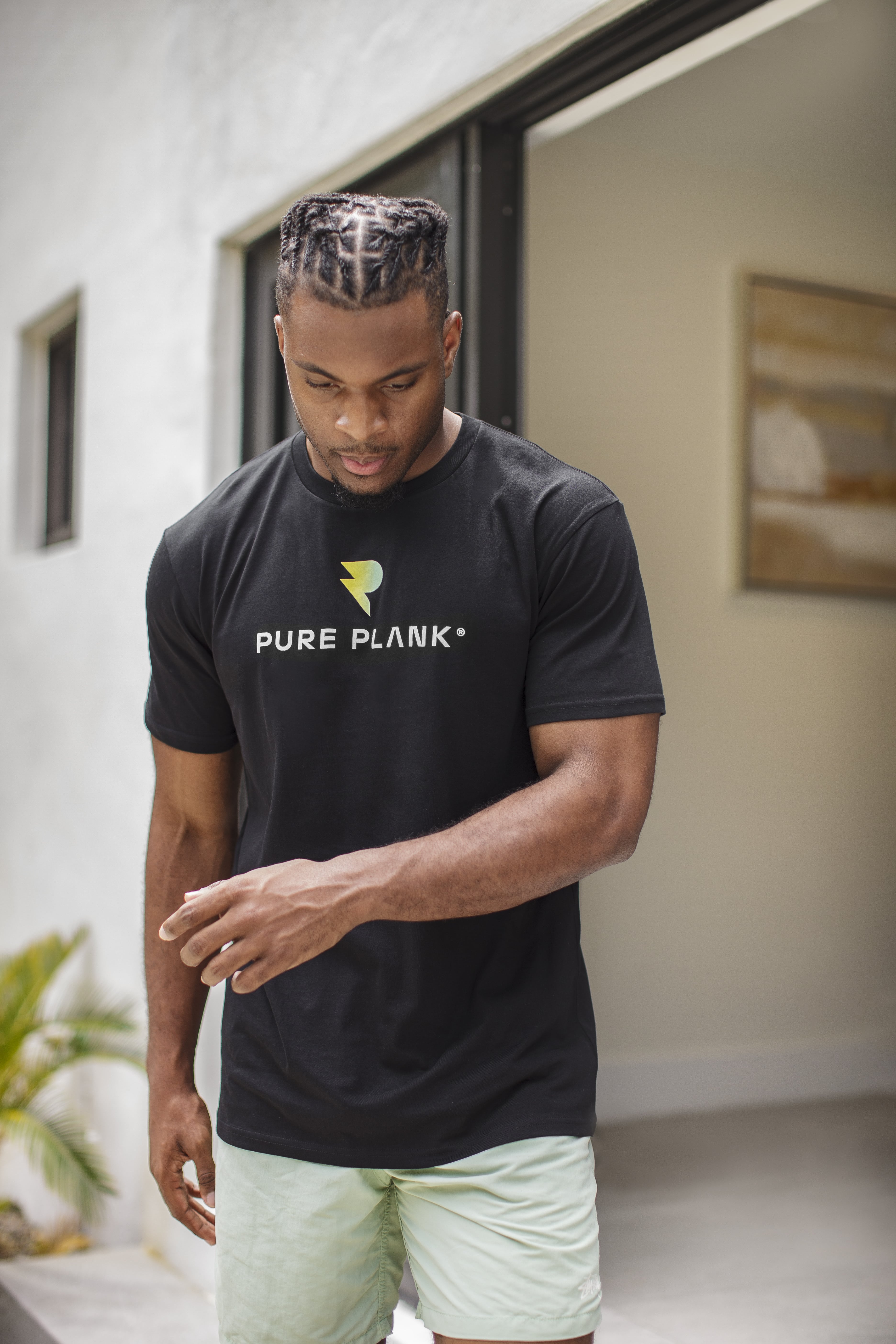 Man in black 'Pure Plank' t-shirt and light shorts, standing outside a building.