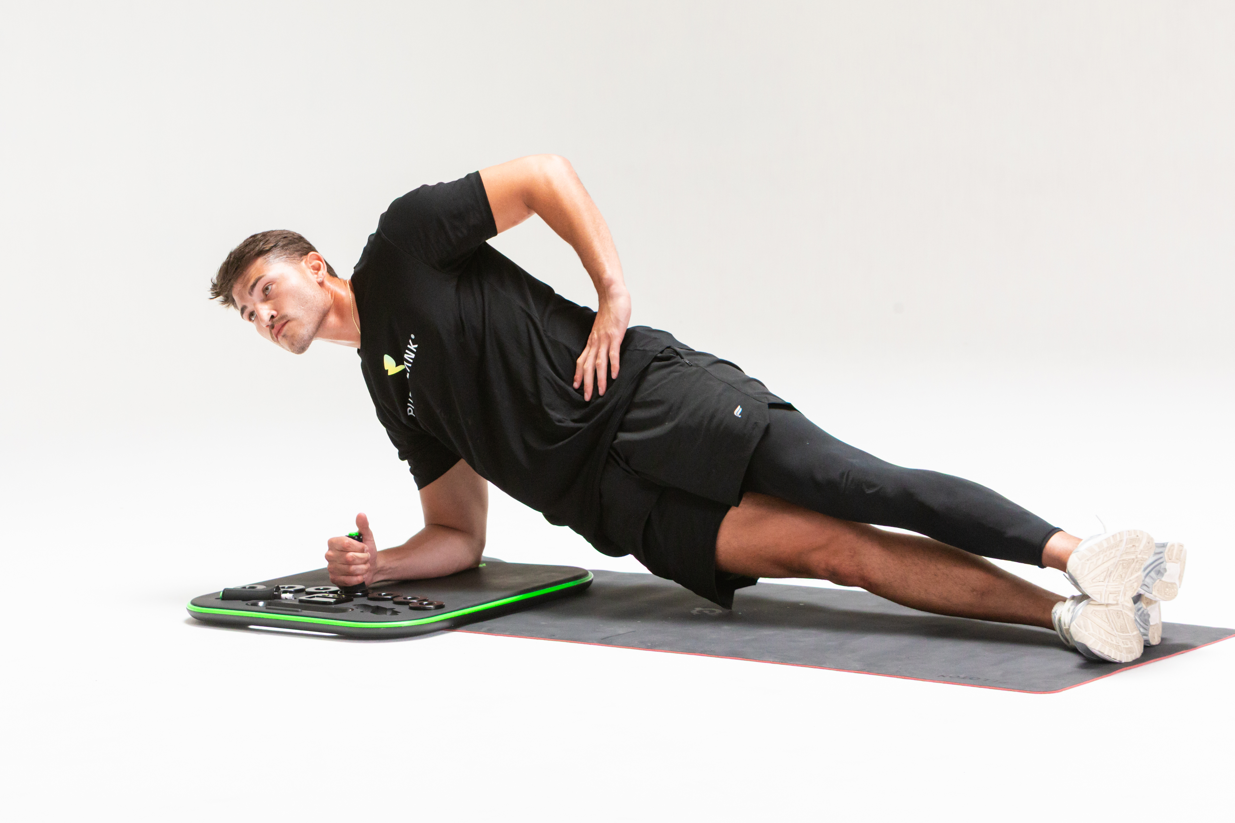 A man in workout clothes holds a side plank on a mat, resting his forearm on an exercise board.