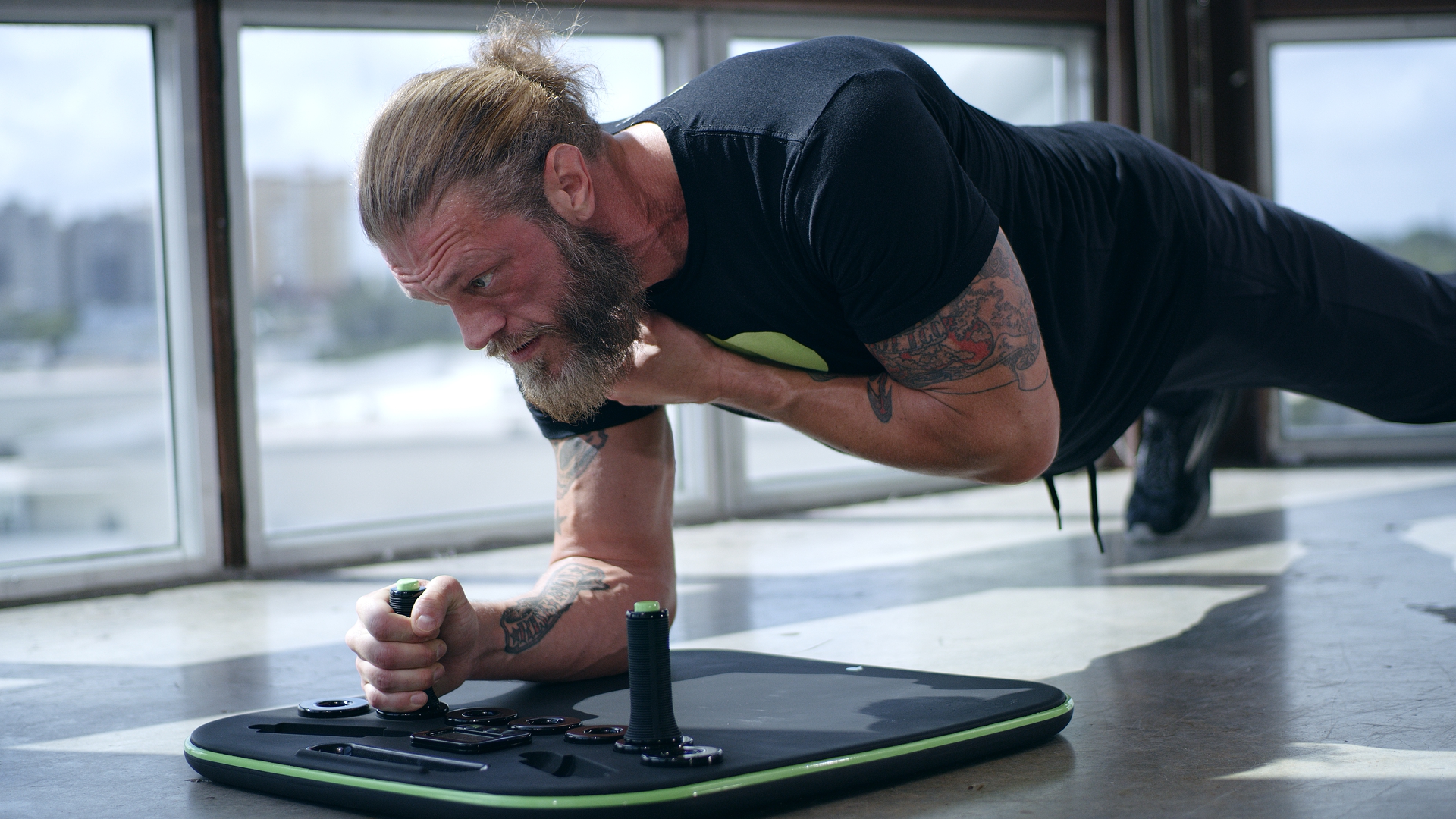 A tattooed man with a beard doing a plank on workout equipment in front of a window.