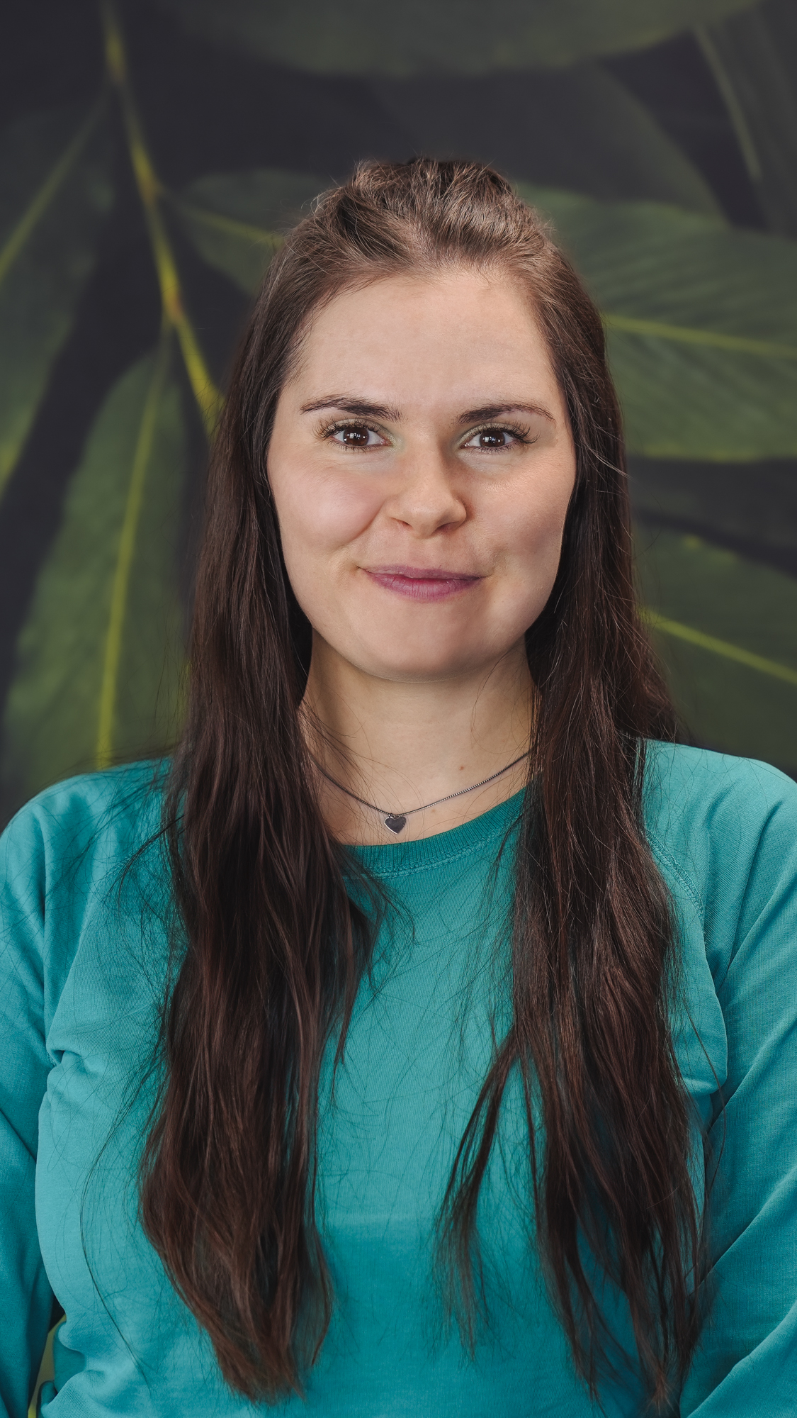 Person with long hair smiling, wearing a teal shirt and necklace, in front of leafy background.