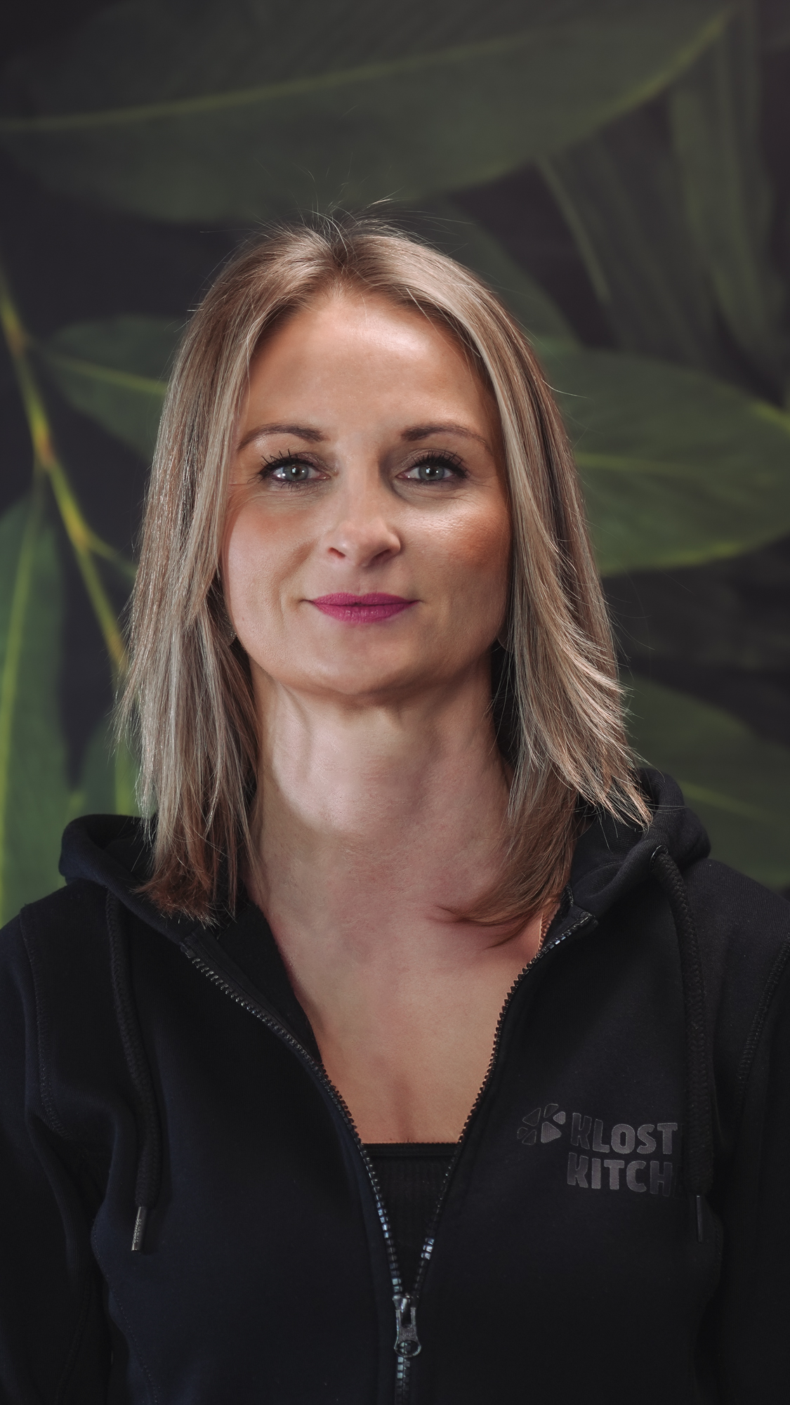 Smiling person in a black hoodie with a leafy background.
