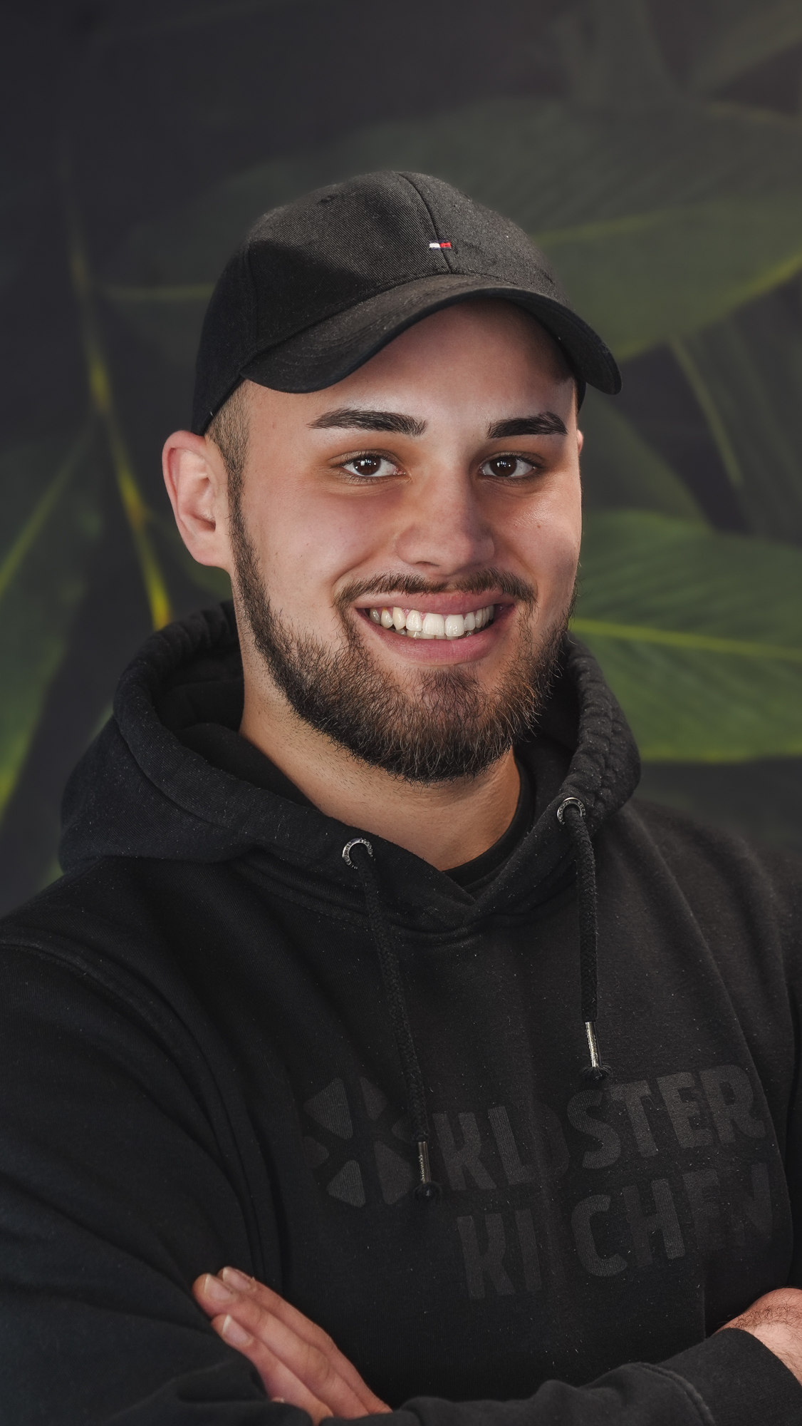 Man in a black hoodie and cap, smiling with arms crossed.