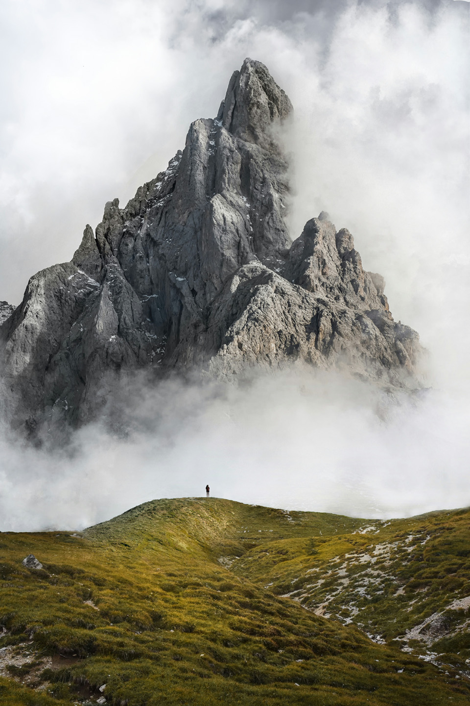 Person standing on grassy hill with towering mountain and clouds.