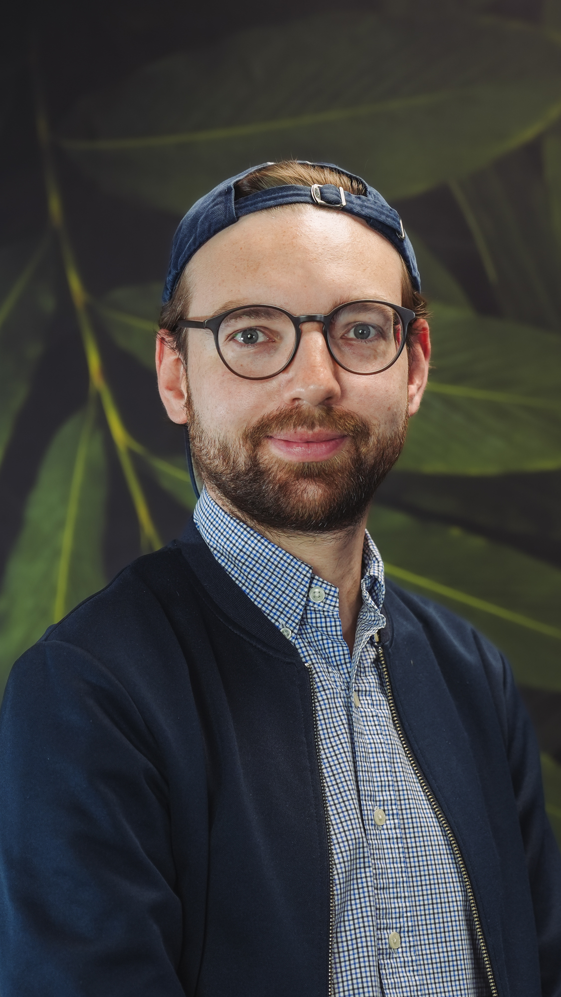 A person wearing glasses and a cap, standing against a leafy background.