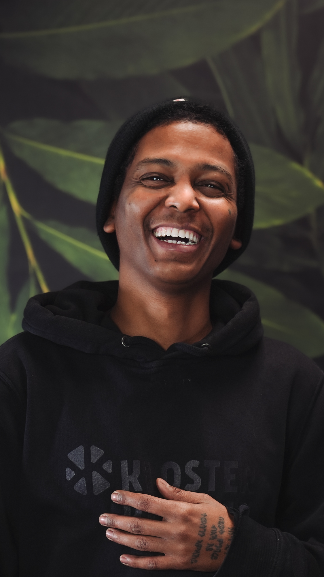 Person in black beanie and hoodie smiling in front of leafy background.