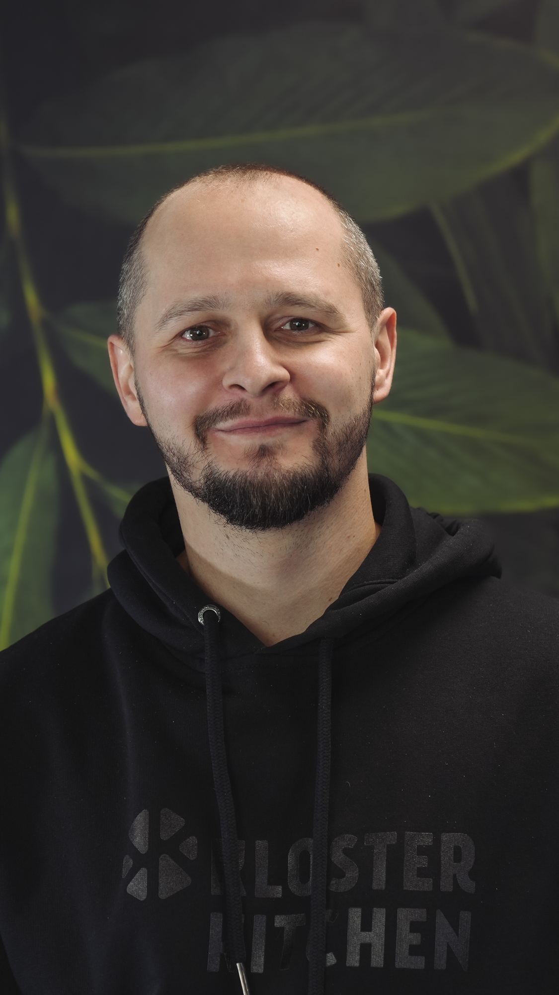 A person in a black hoodie smiles against a leafy background.