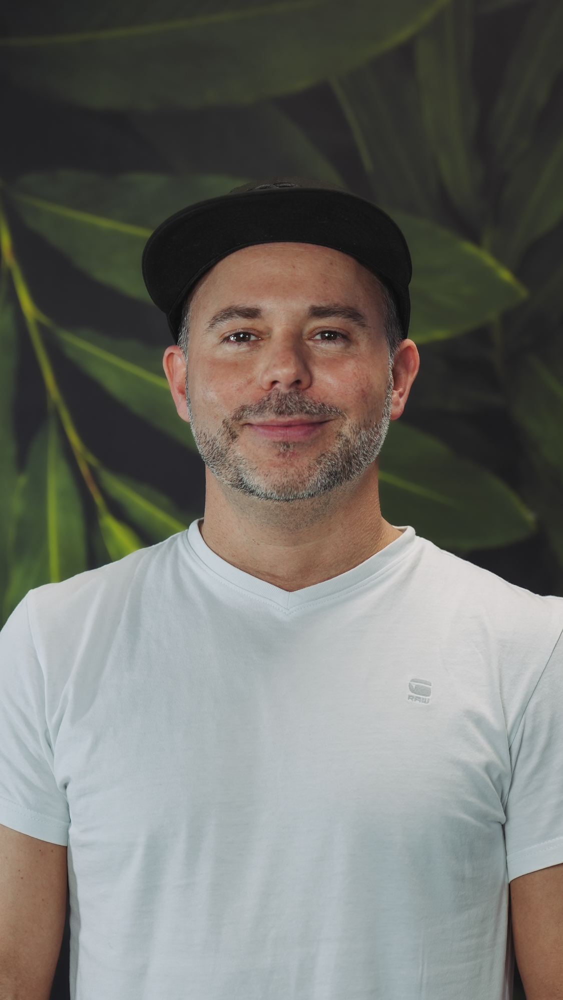 A person wearing a cap and white shirt, smiling with a leafy background.