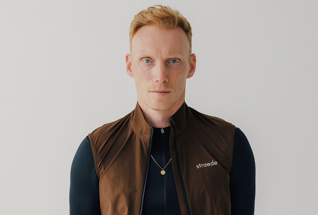 Un hombre pelirrojo y de ojos azules que lleva un chaleco marrón sobre una camisa oscura, mirando hacia delante.