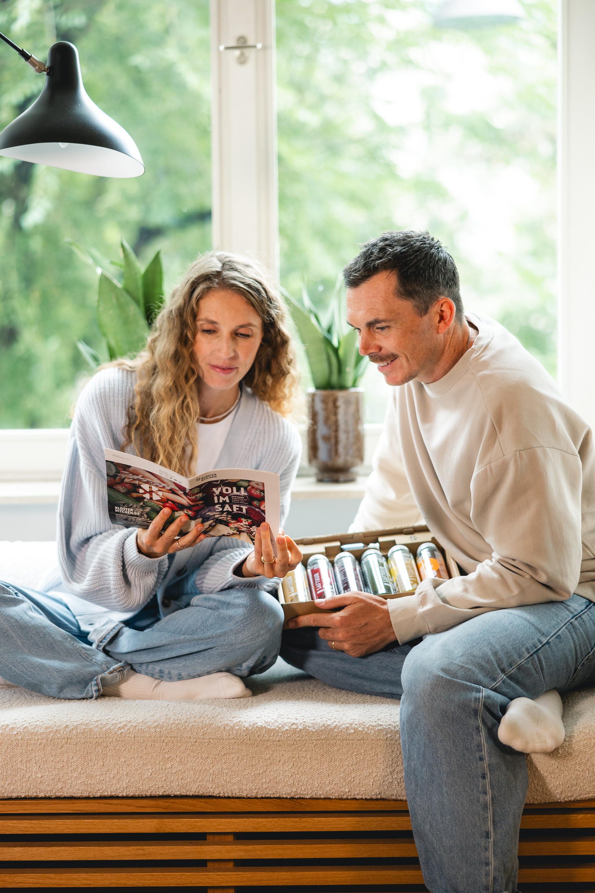 Dos personas sentadas junto a una ventana, leyendo un folleto y sosteniendo una caja de bebidas.
