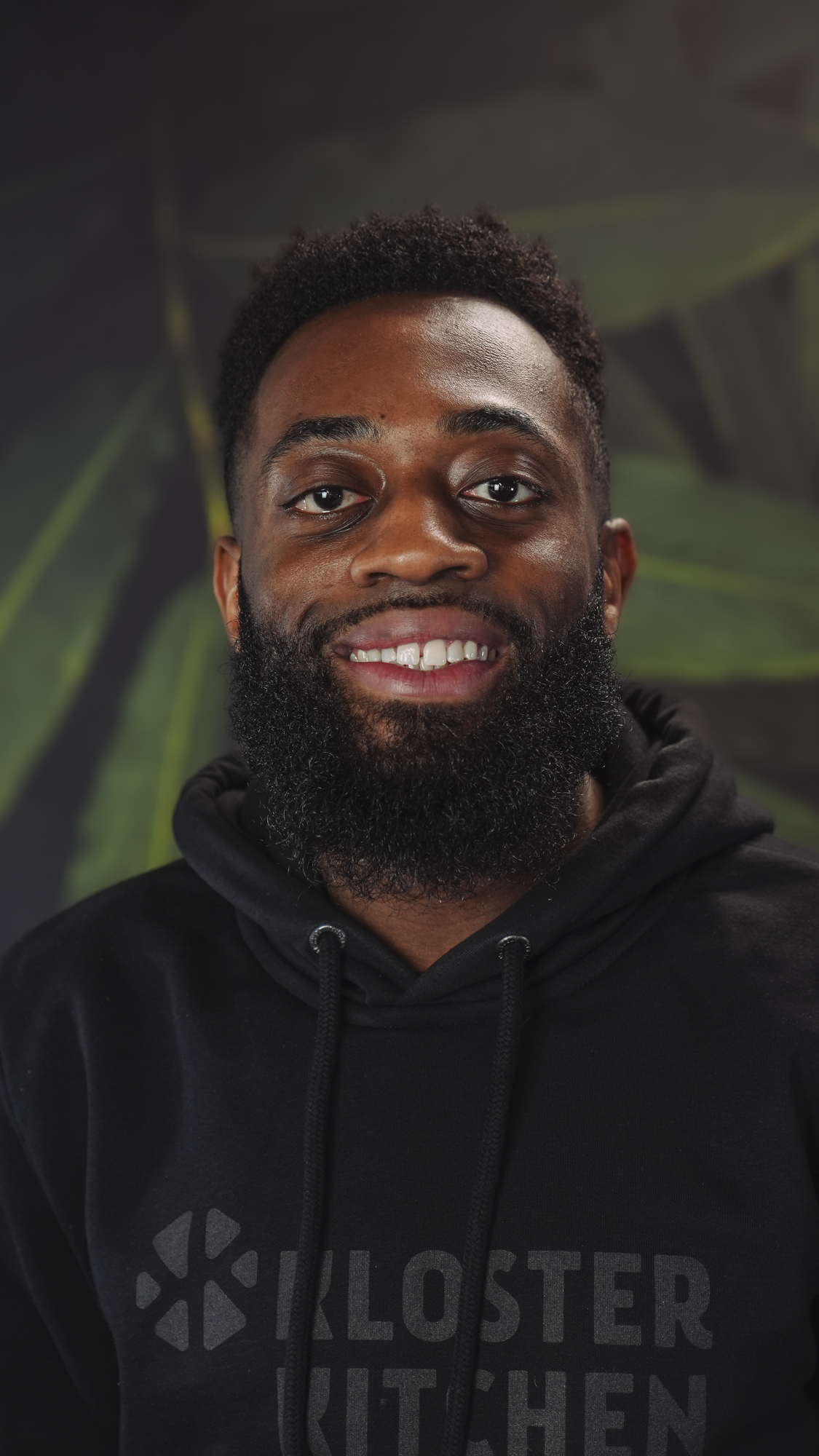 Man smiling, wearing a black hoodie withKLOSTER KITCHEN' logo, leafy background.