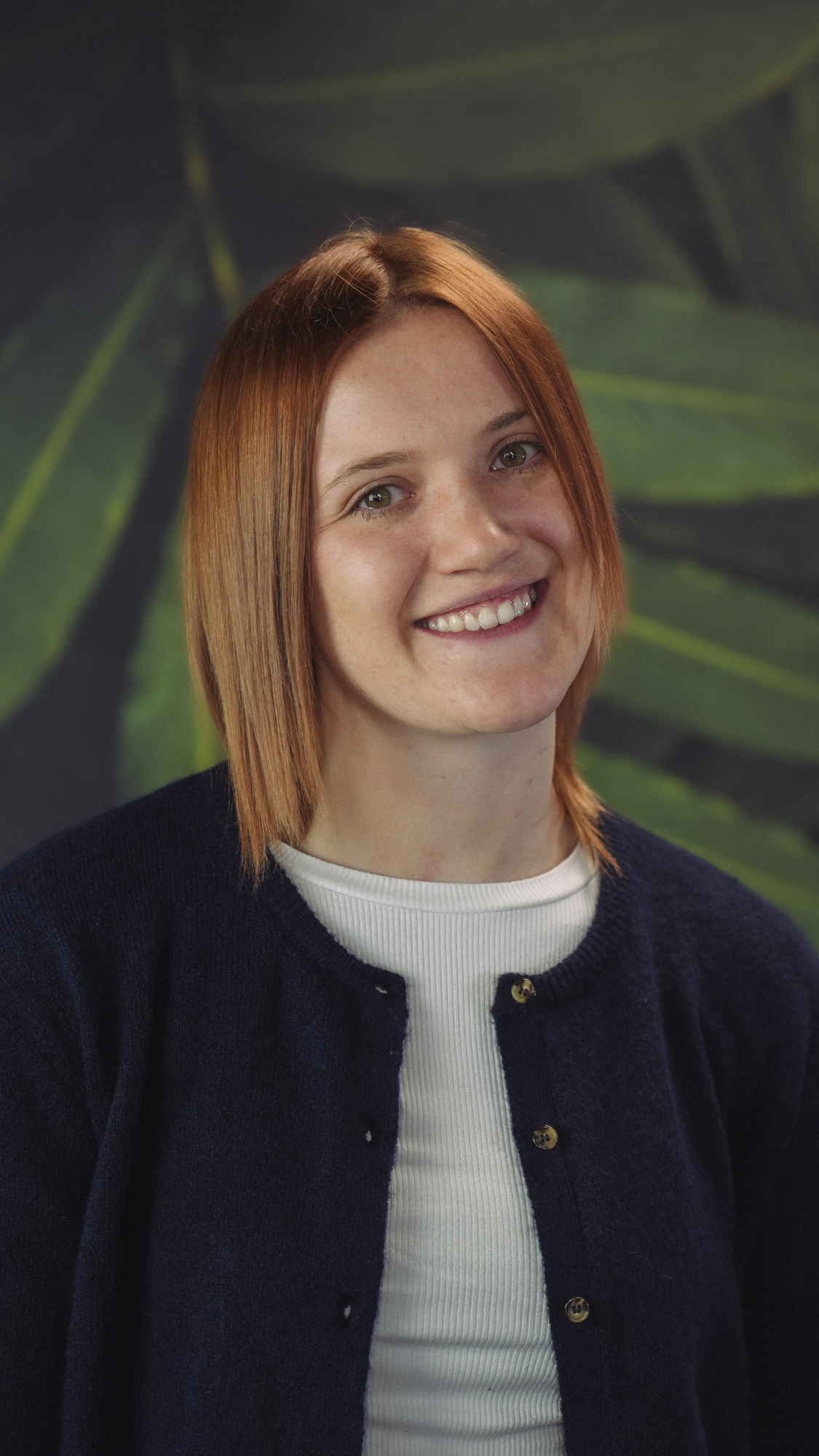 Person with reddish hair wearing a black hoodie, smiling in front of a leafy background.