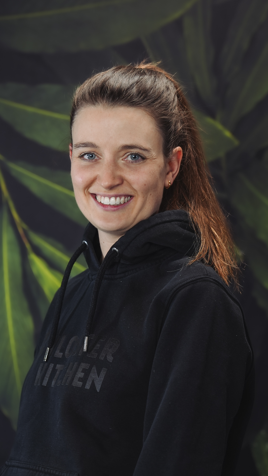 A person smiling in front of a leafy background wearing a black hoodie.
