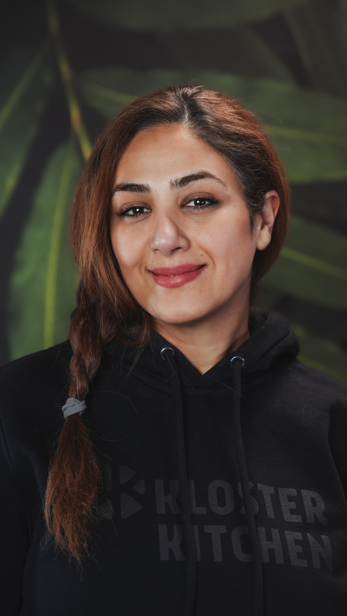 Person in a black hoodie with a braided hairstyle, smiling in front of a leafy background.