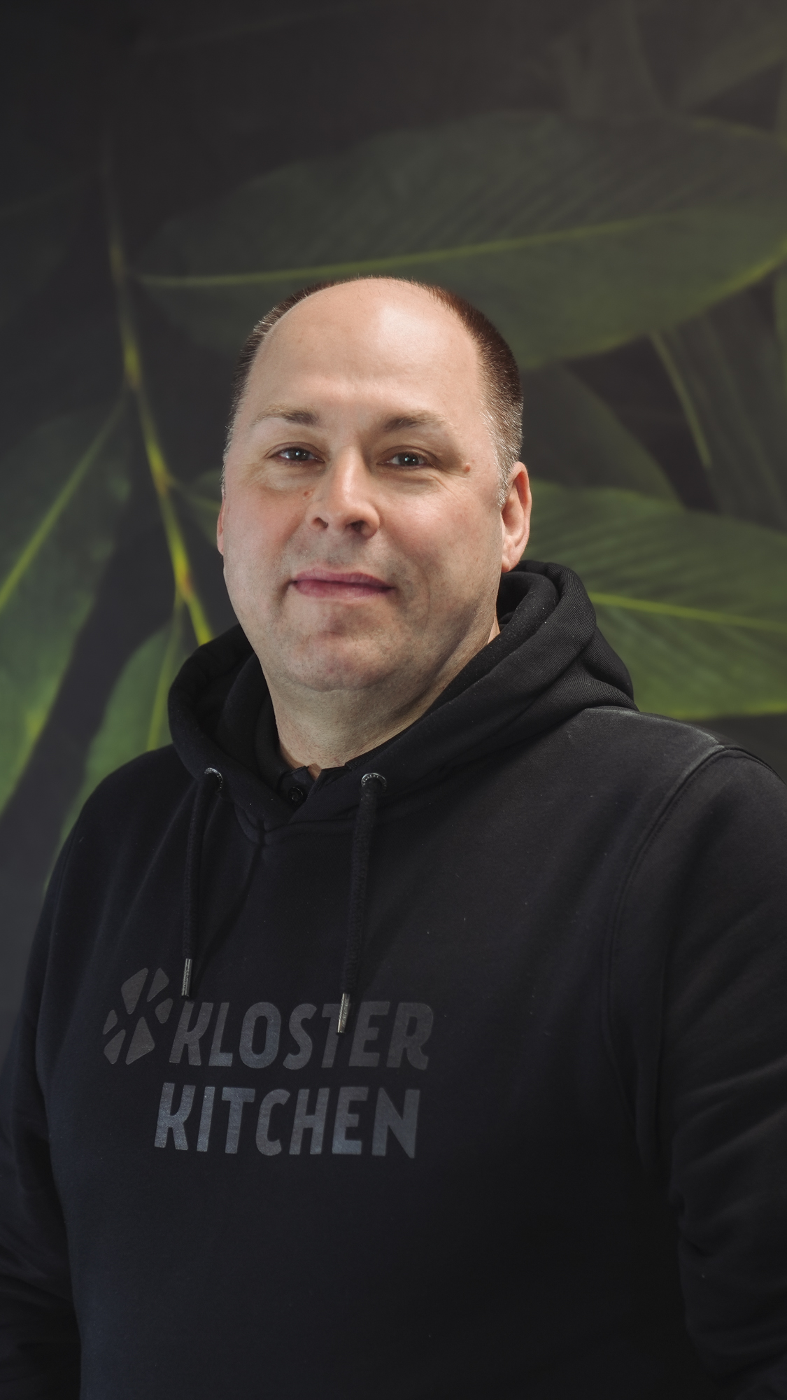 A person wearing a white beanie and black hoodie, smiling against a leafy background.