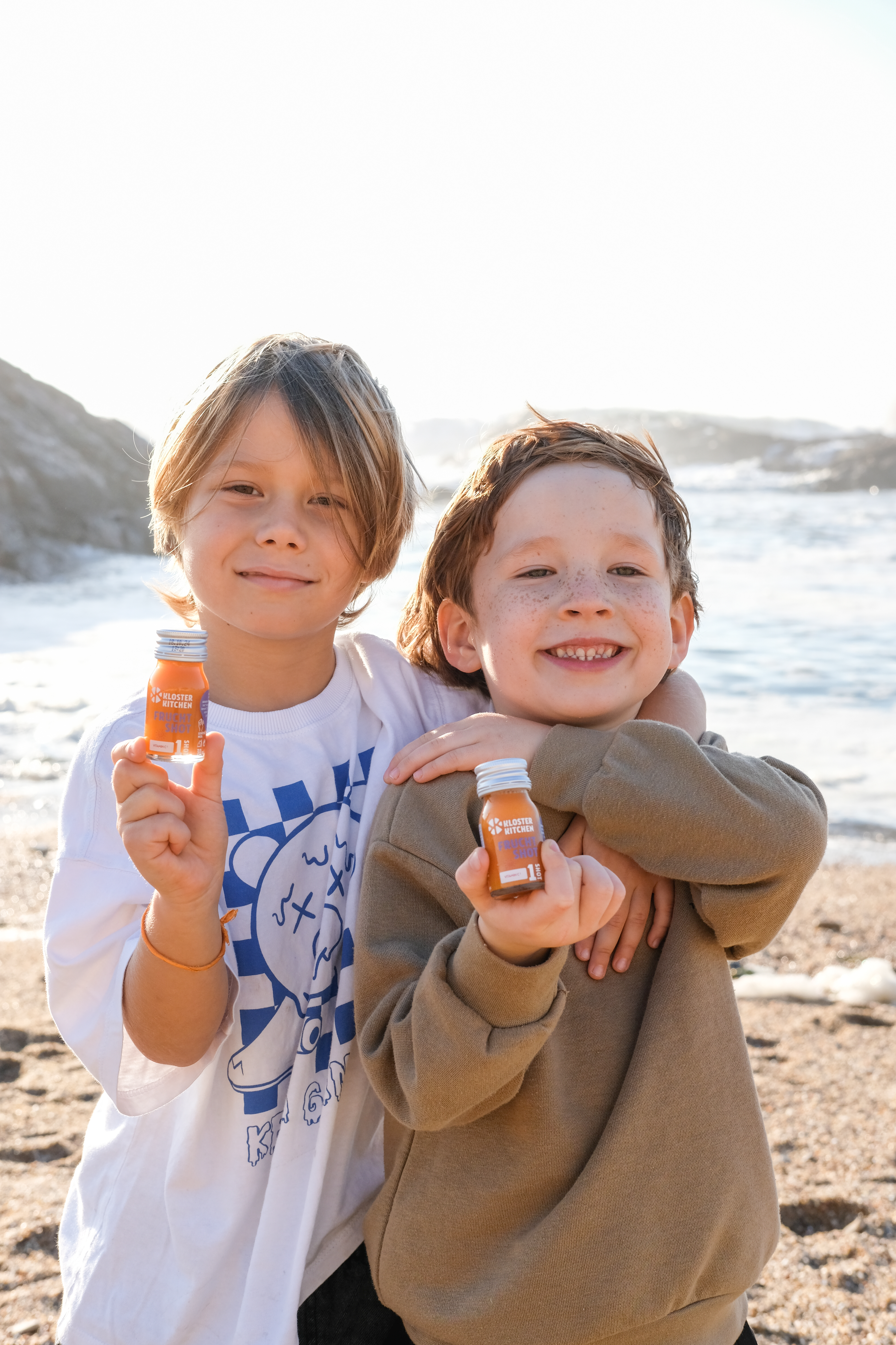 Two children holding small bottles on a beach, smiling at the camera.