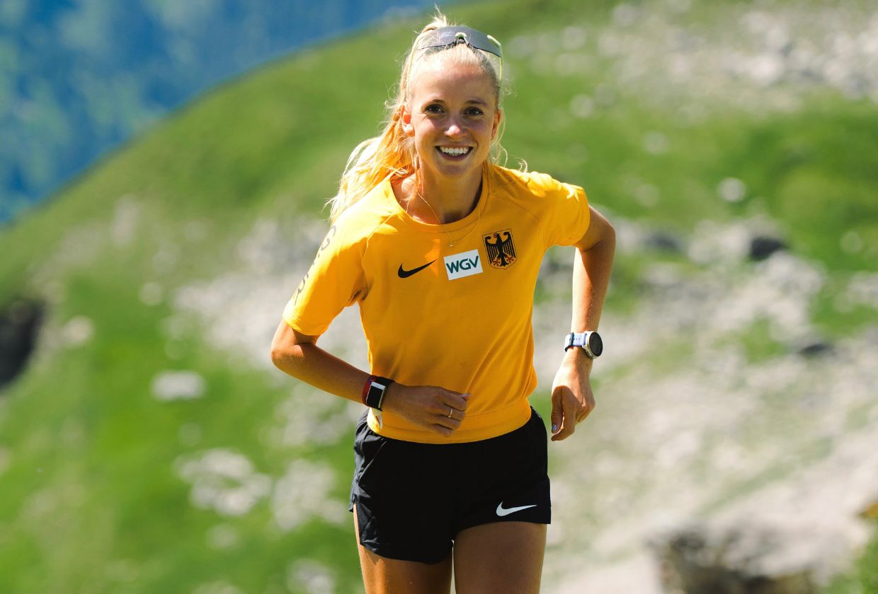 Una donna bionda sorridente con una maglietta gialla e pantaloncini neri corre su una collina erbosa.