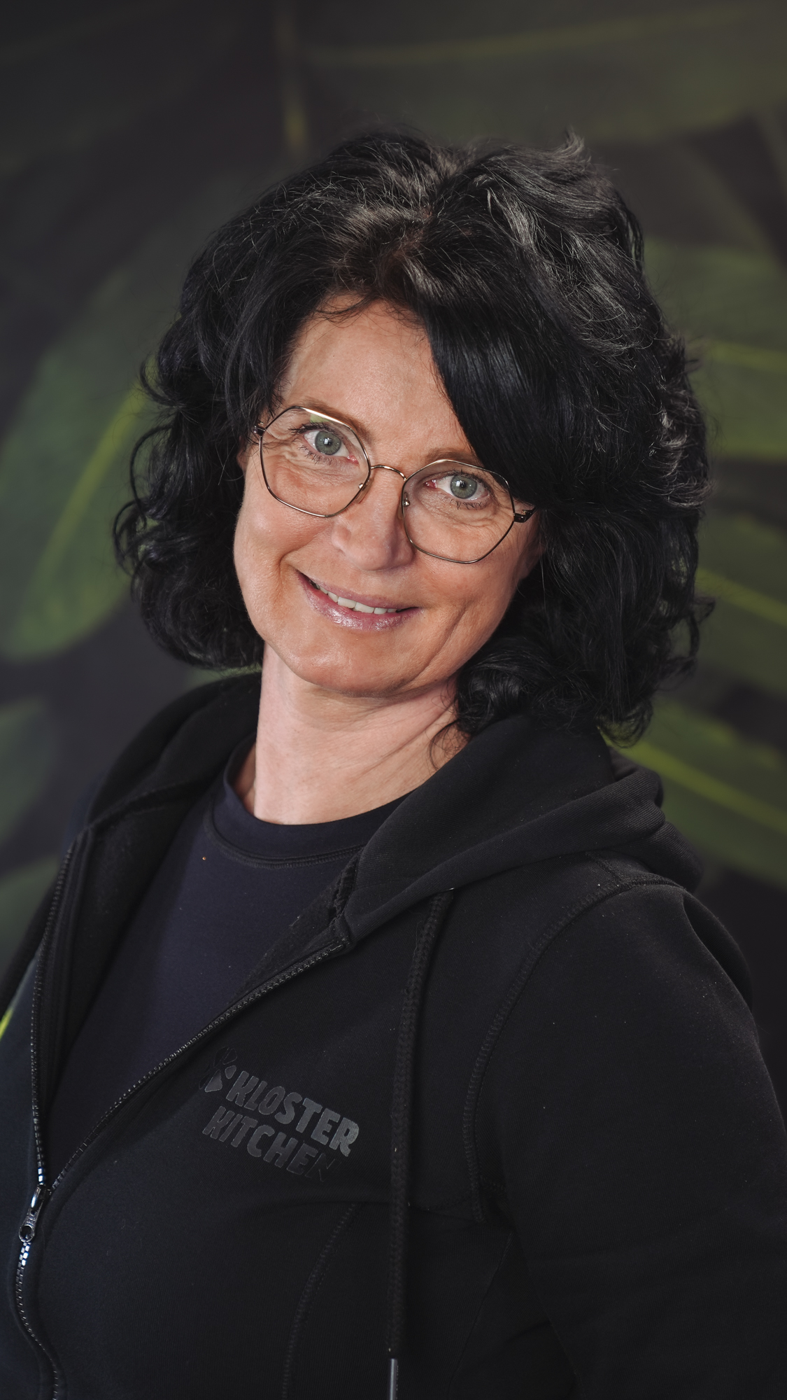 A woman with glasses smiles, wearing a black hoodie against a leafy background.