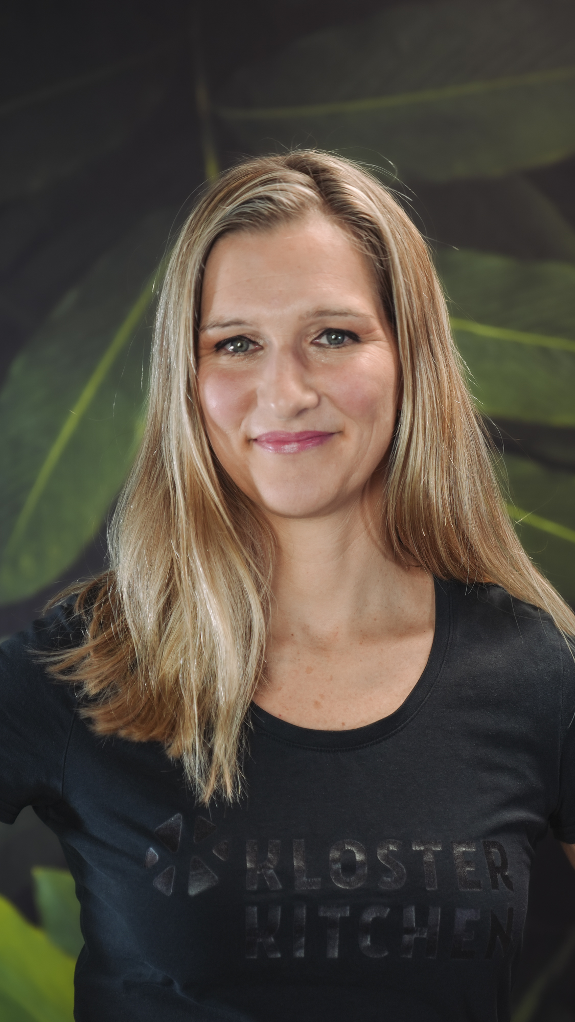 A person with long hair smiling, wearing aKloster Kitchen' shirt.