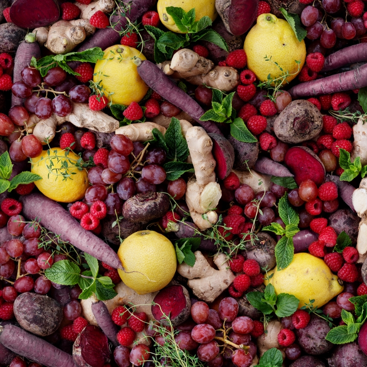 A flat lay of colorful fresh produce including lemons, raspberries, grapes, ginger, beets, and purple carrots.