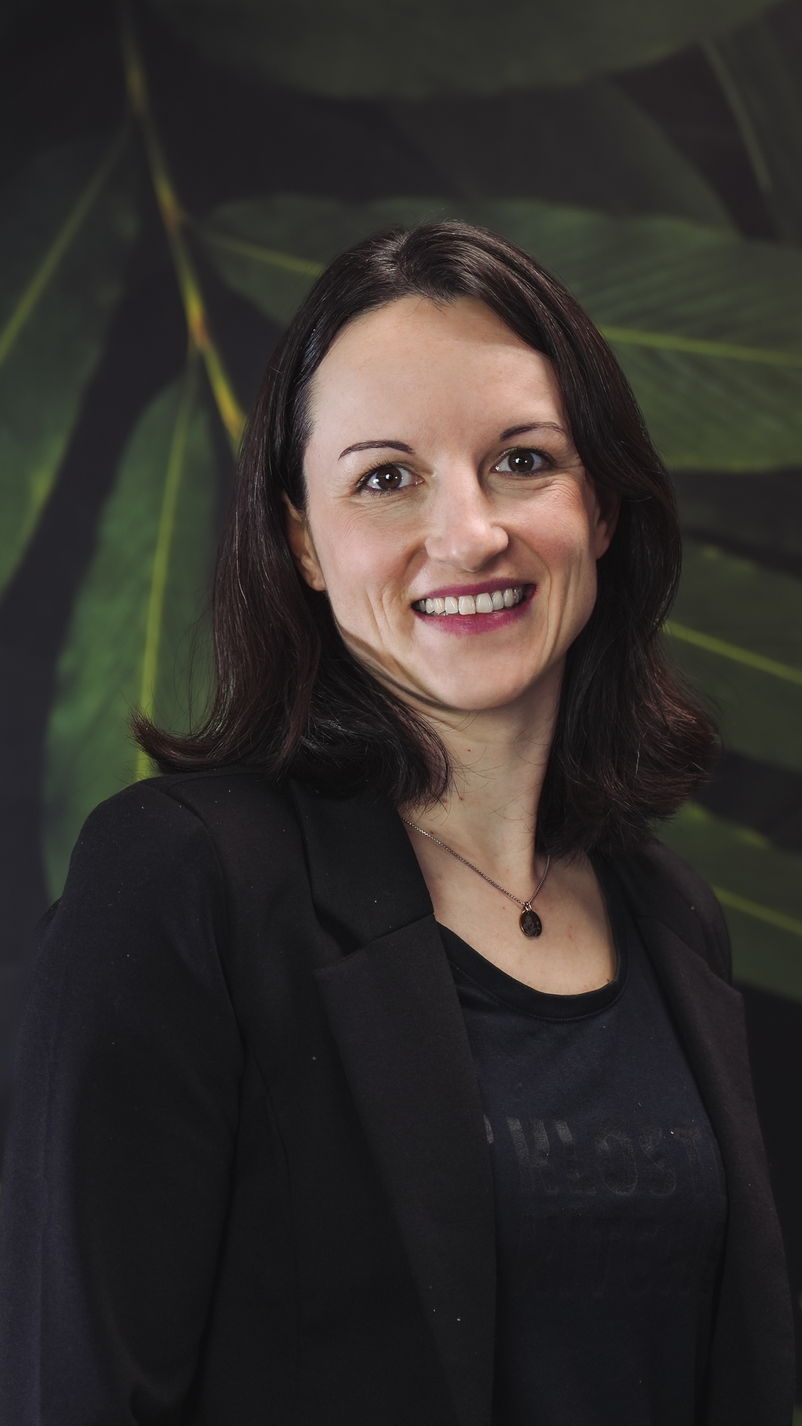 A woman smiling, wearing a black blazer with a leafy background.