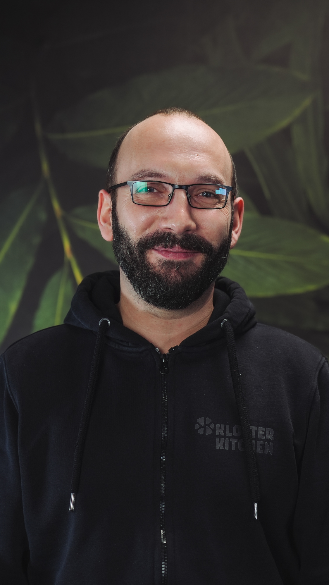 A person wearing glasses and a hoodie, smiling in front of leafy background.